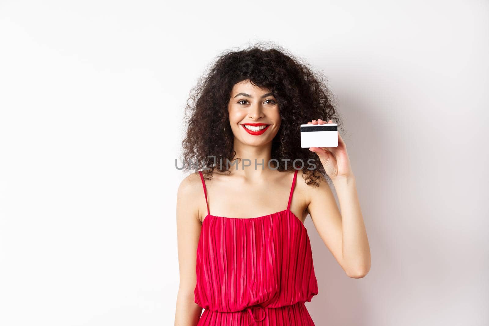 Beautiful woman in red trendy dress and makeup, showing plastic credit card and smiling, recommending offer, standing on white background.