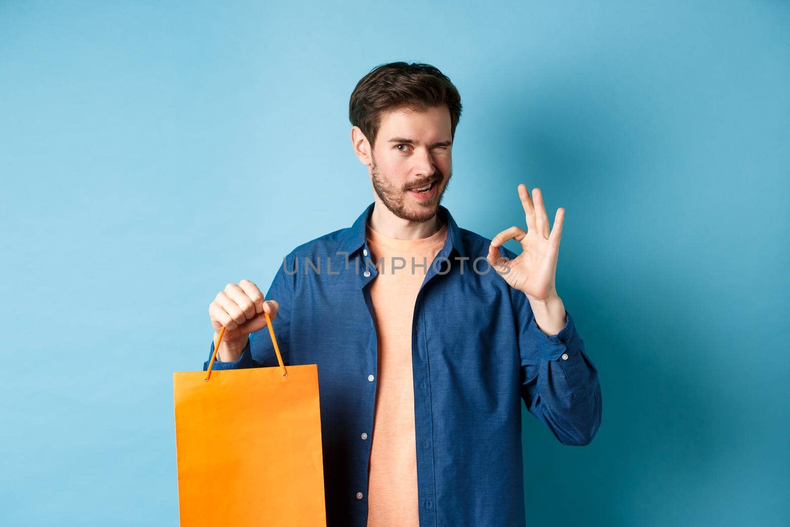 Modern man in casual outfit showing shopping bag and okay sign, winking at camera, recommending shop, standing on blue background by Benzoix