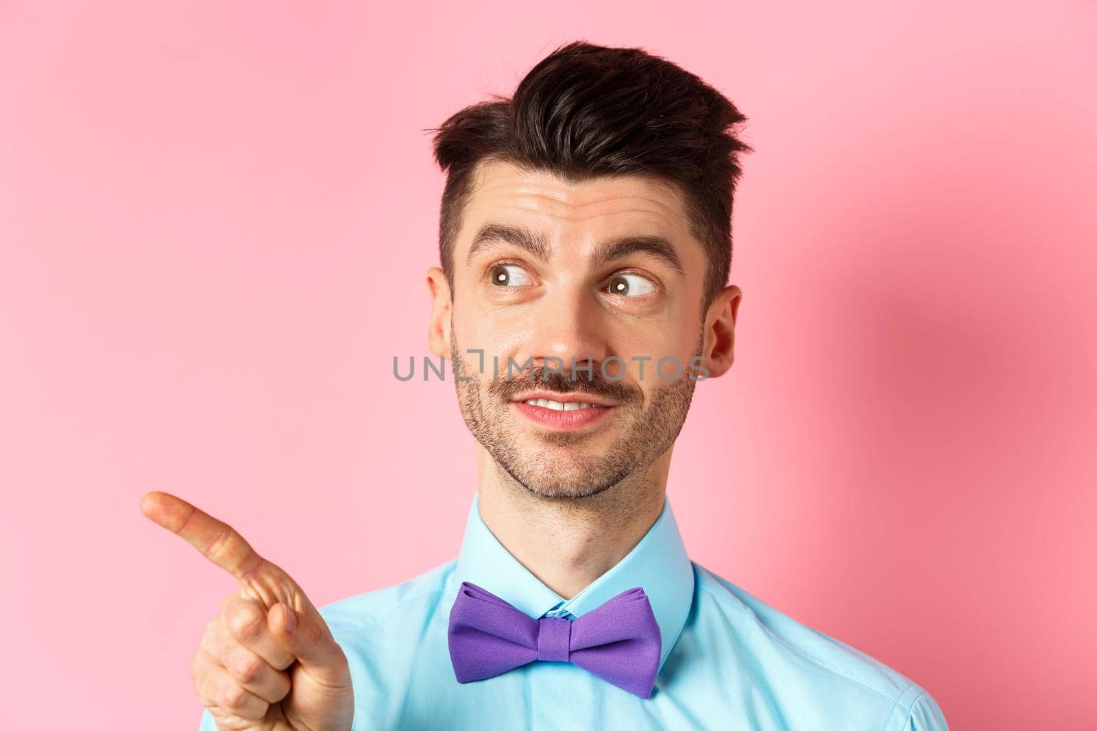 Close-up of caucasian male model with moustache and bow-tie, looking and pointing left with intrigued smile, showing promo offer, standing in bow-tie on pink background.