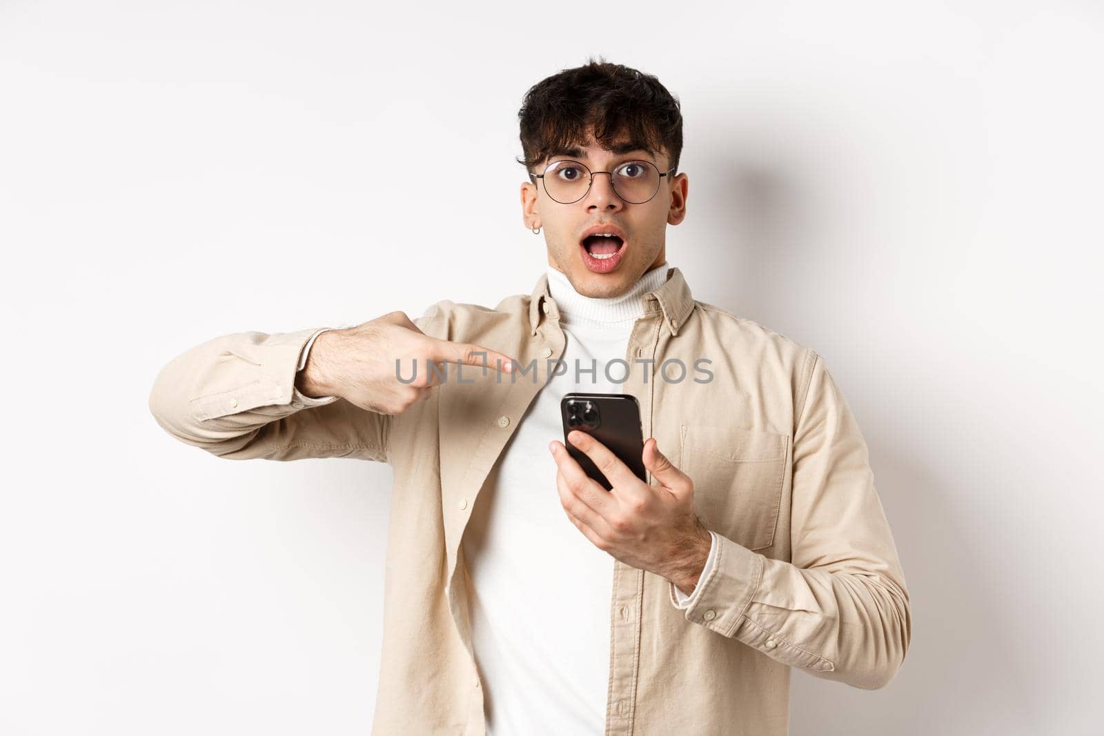 Real people. Amazed young guy checking out online offer on mobile phone, pointing at smartphone screen with excited face, standing on white background.