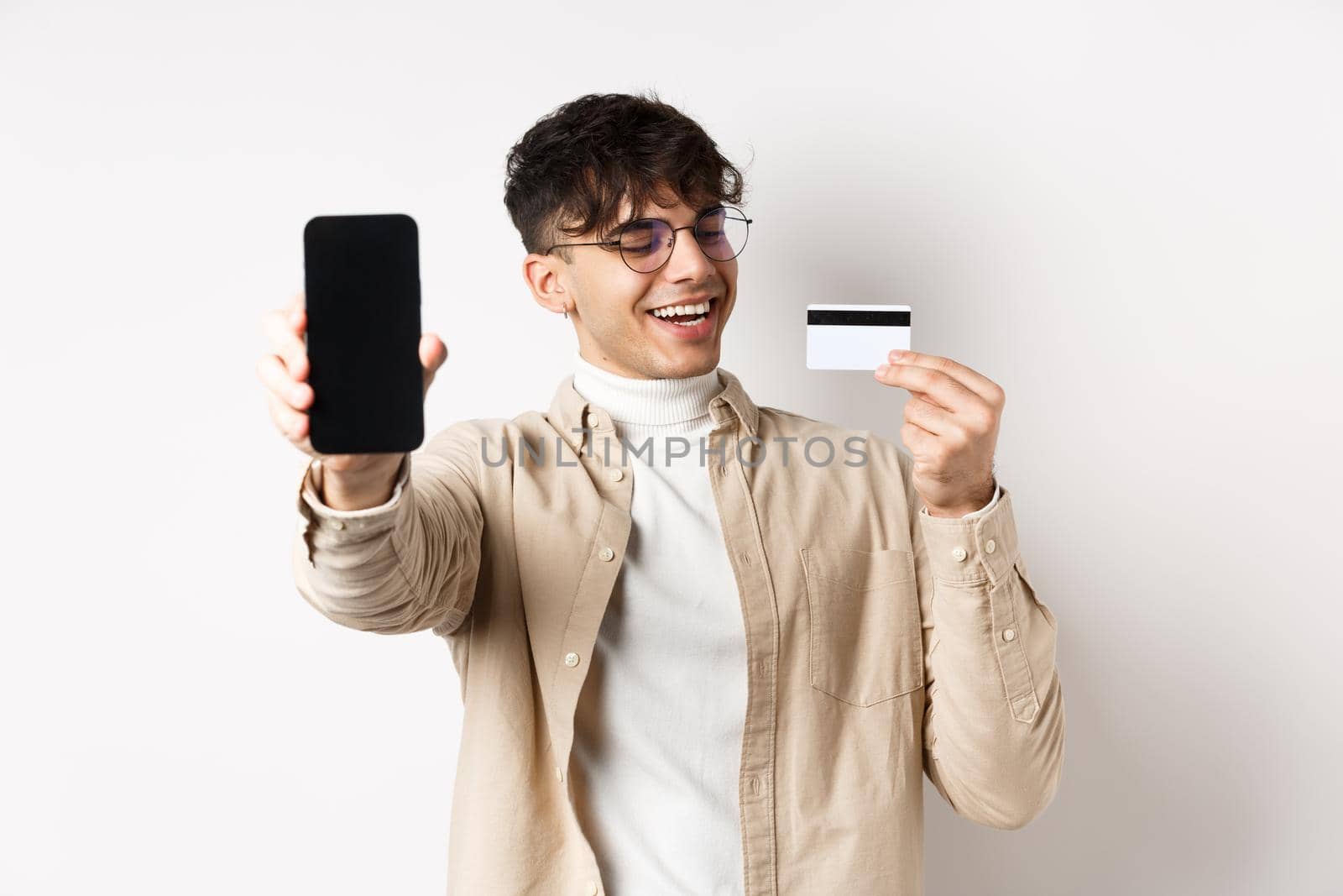 E-commerce. Happy young man looking satisfied at plastic credit card, showing smartphone screen to boast, standing on white background.