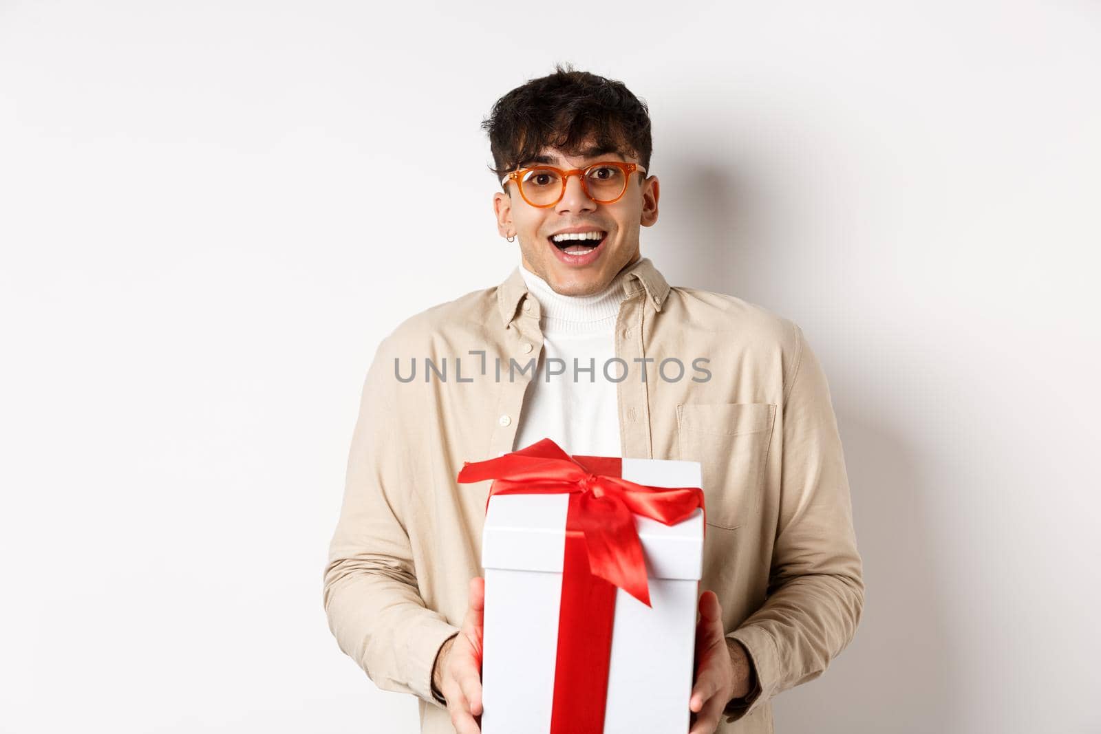 Surprised happy guy receiving a gift, holding present box and smiling amazed, standing on white background.