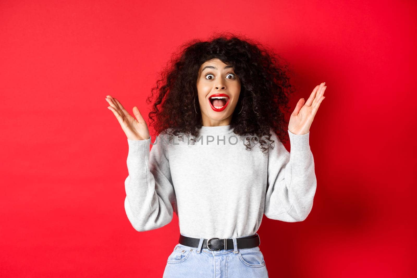 Image of happy and surprised curly woman in makeup and sweatshirt, raising hands up and rejoicing from good news, standing on red background.