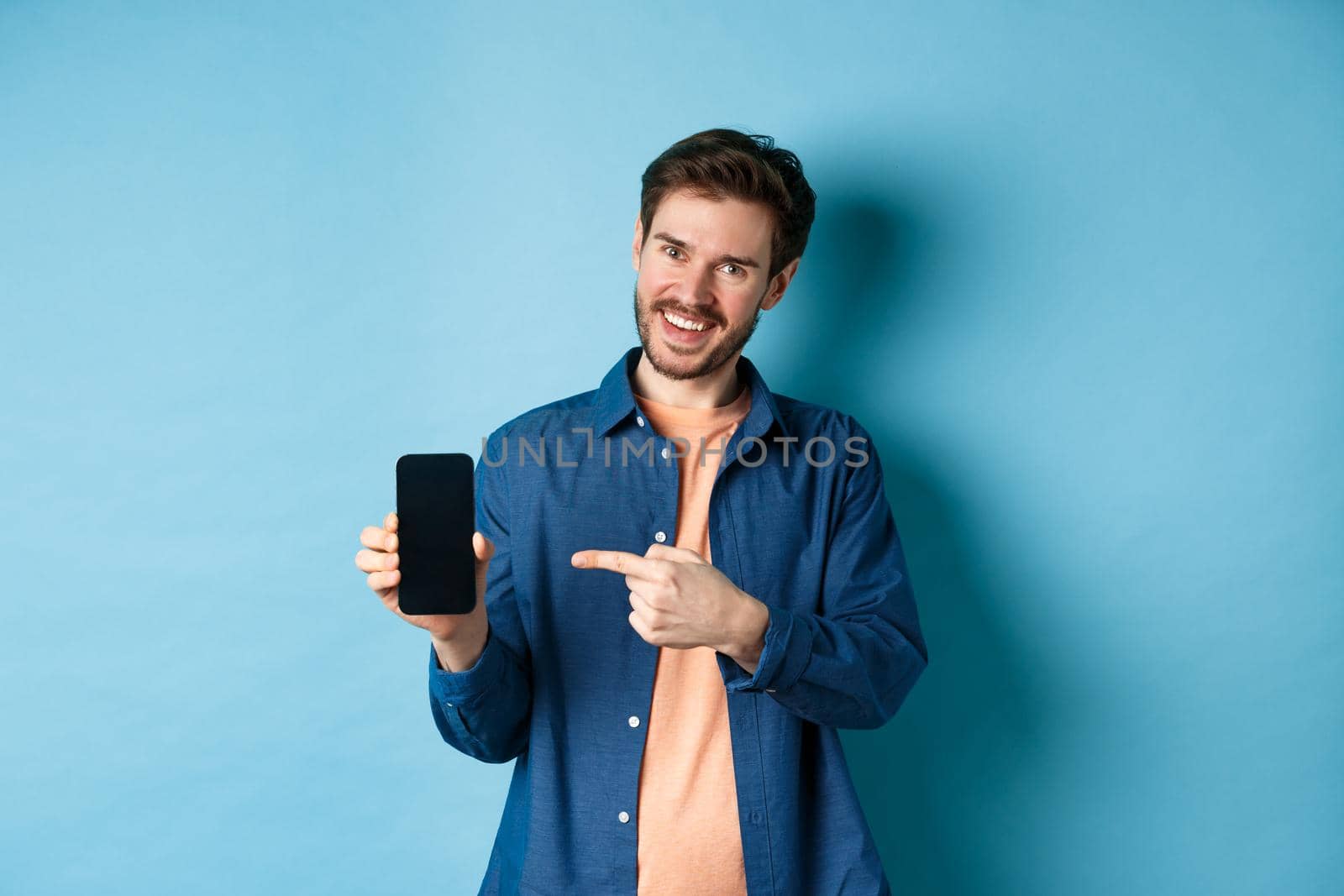 Smiling caucasian man showing on empty smartphone screen, pointing at mobile phone and looking satisfied, standing on blue background by Benzoix