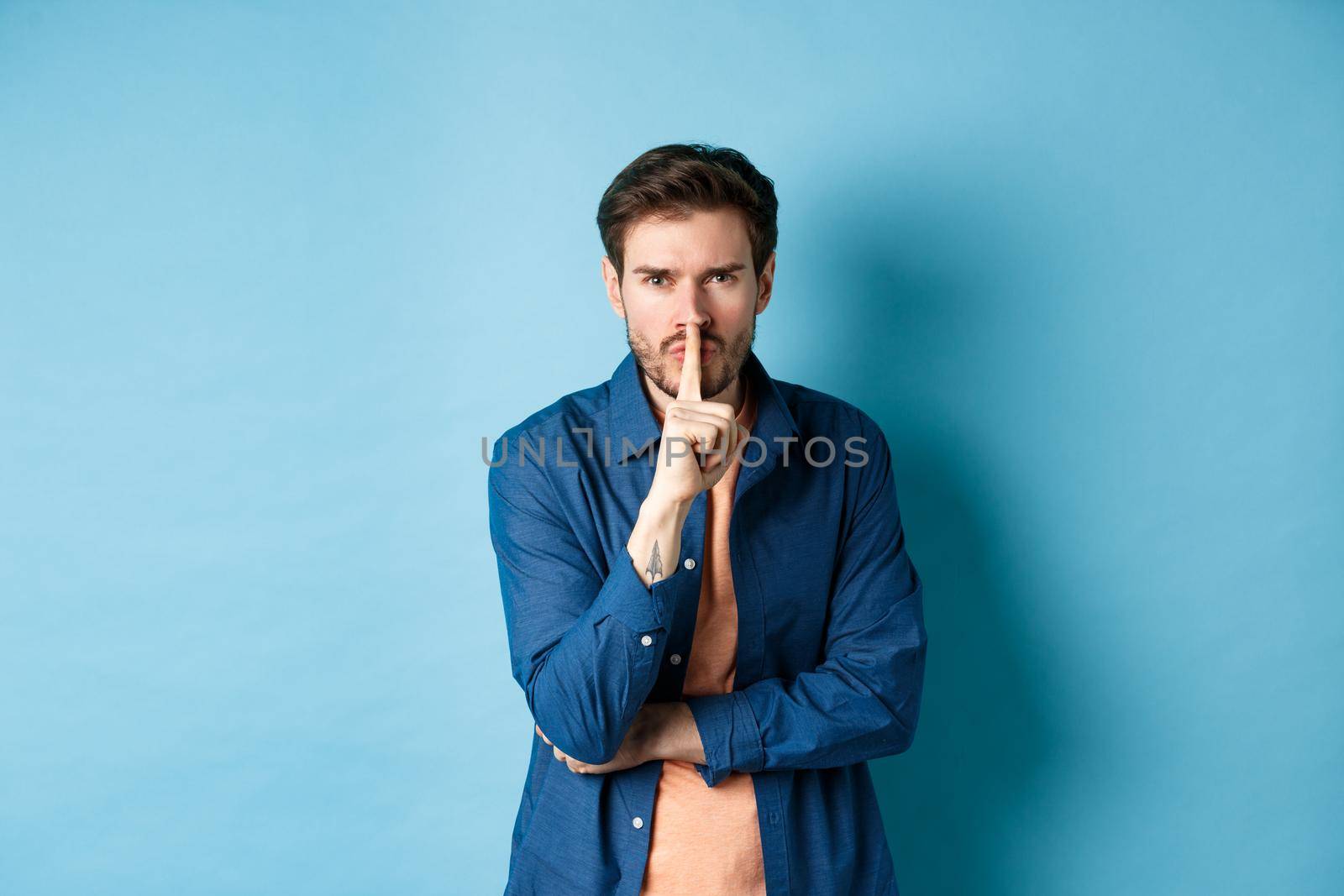 Angry man shushing, press finger to lips and frowning, tell to be quiet, show taboo gesture, standing on blue background.