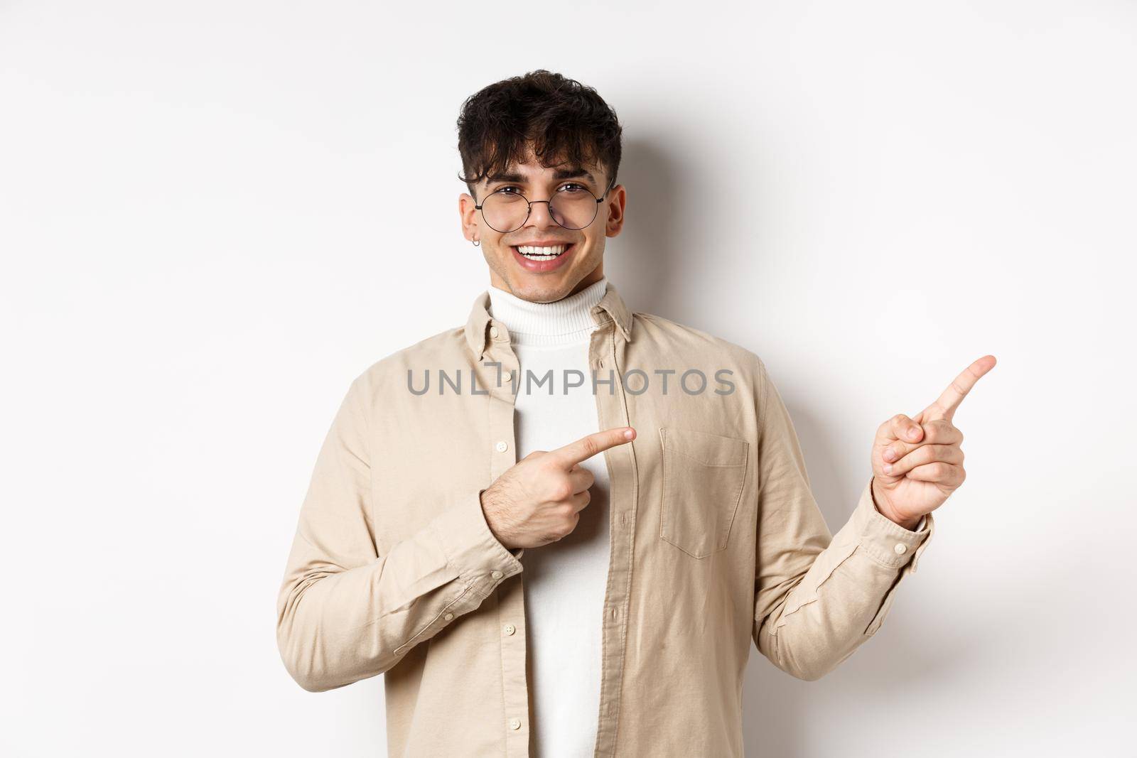 Proud and happy young man in glasses showing logo, pointing fingers at upper right corner and smiling, standing on white background.