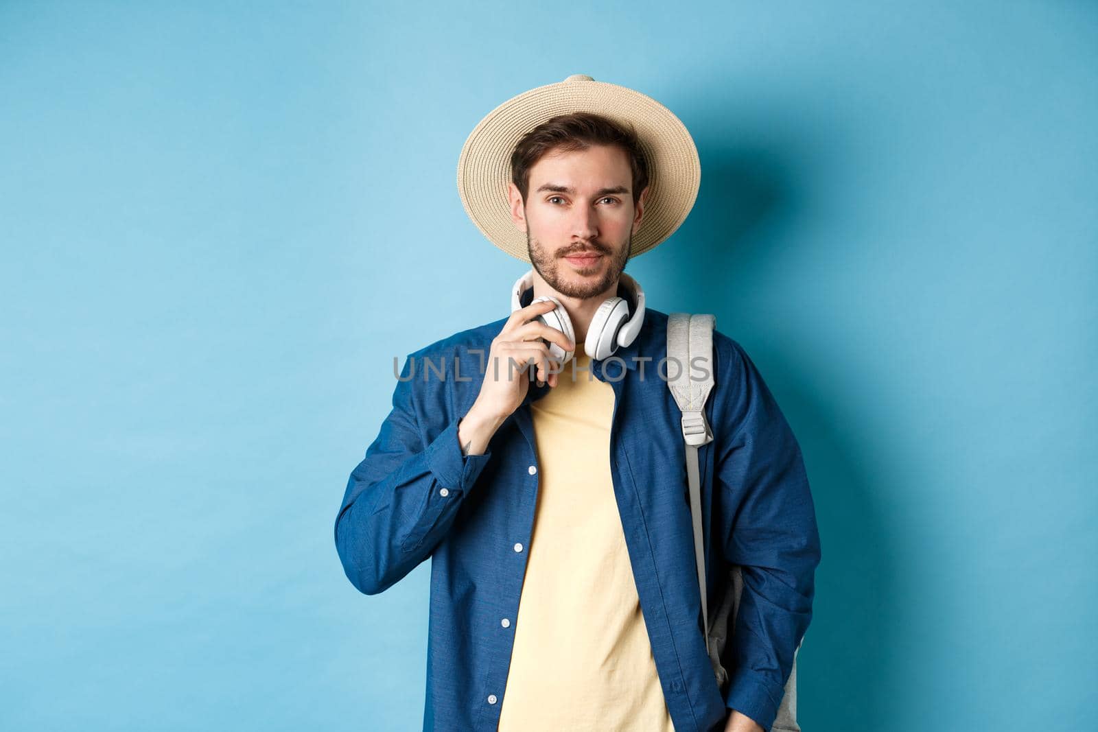 Handsome guy travelling with headphone, wearing straw hat and smiling, standing with backpack on blue background.