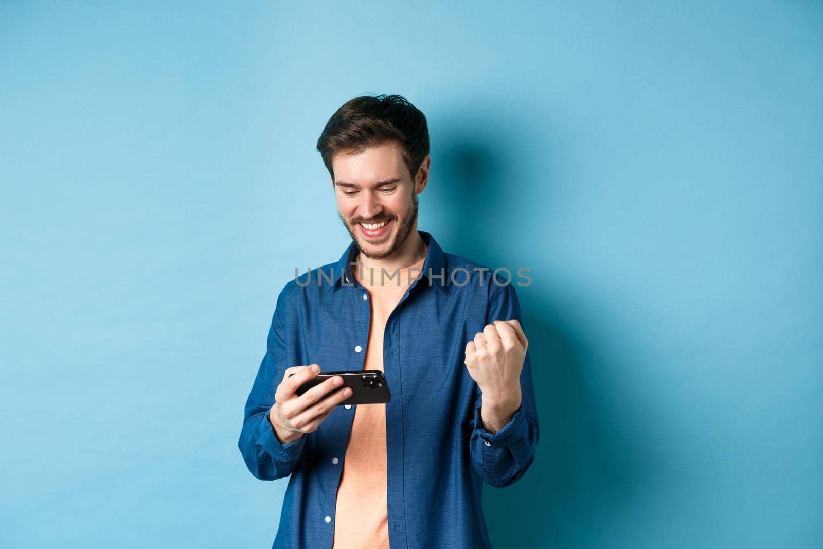 Happy young man winning in video game on mobile phone, looking satisfied at smartphone screen and say yes with joy, standing on blue background.