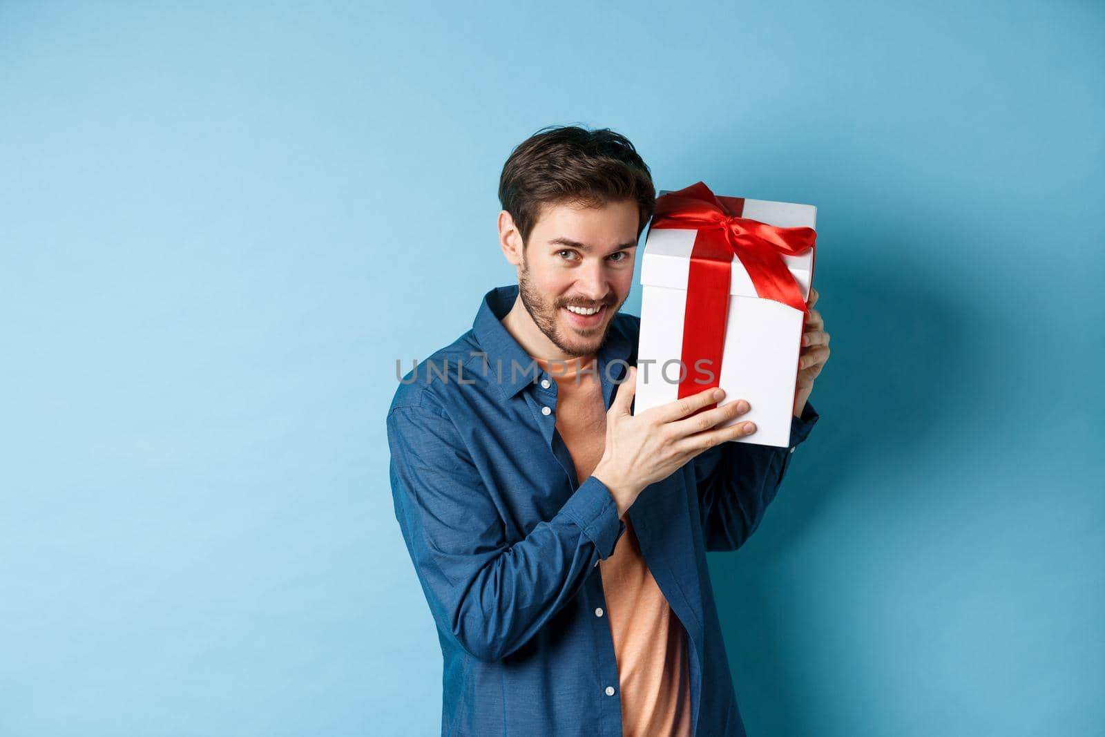 Valentines day. Romantic boyfriend showing gift box for lover, celebrating anniversary, standing over blue background.