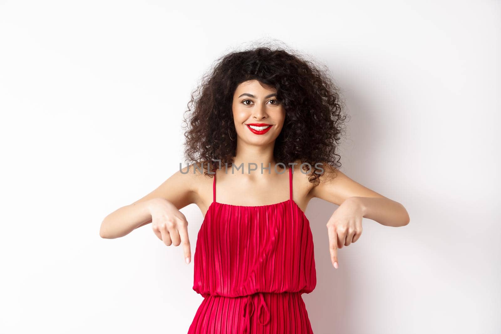 Attractive female model in red dress and makeup, pointing fingers down and smiling, showing advertisement, standing over white background.