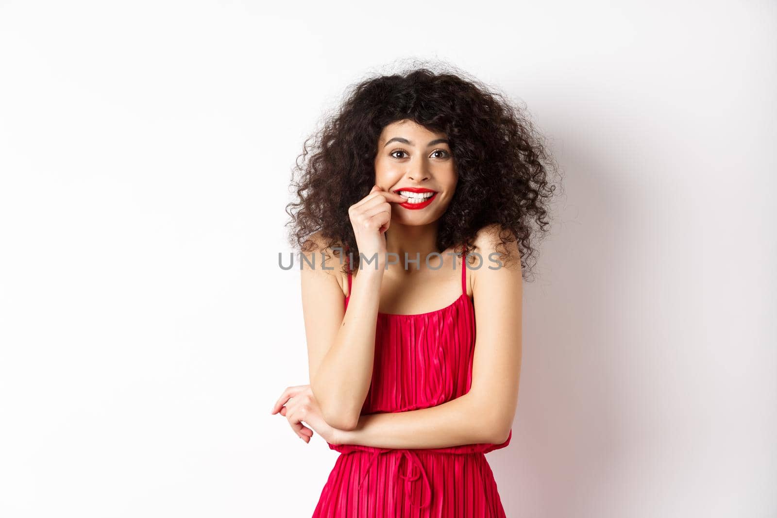 Intrigued young woman with curly hair, wearing elegant red dress and lipstick, biting fingernails and looking interested, standing over white background.