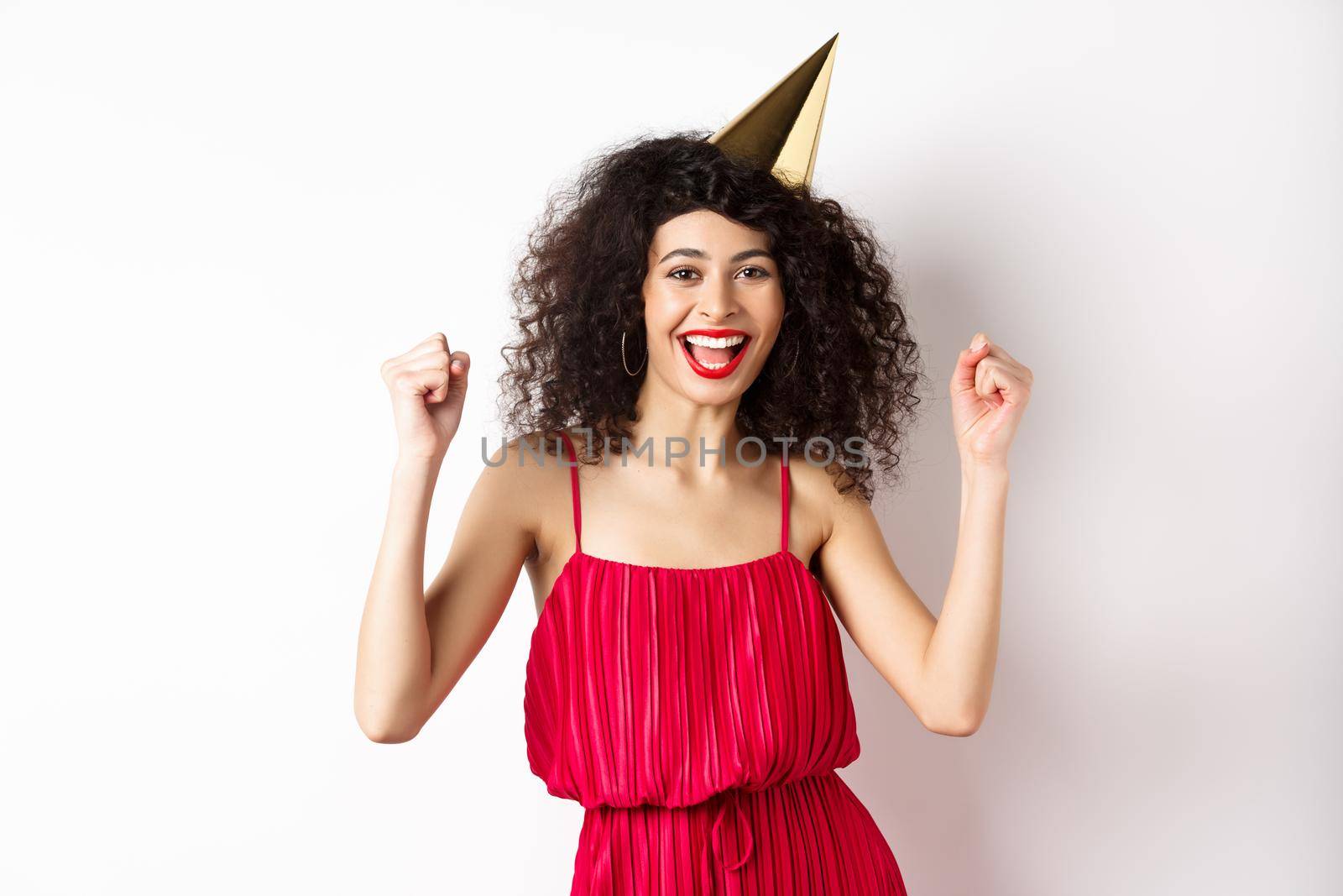 Birthday girl in party hat having fun, dancing in red dress and chanting, standing against white background.