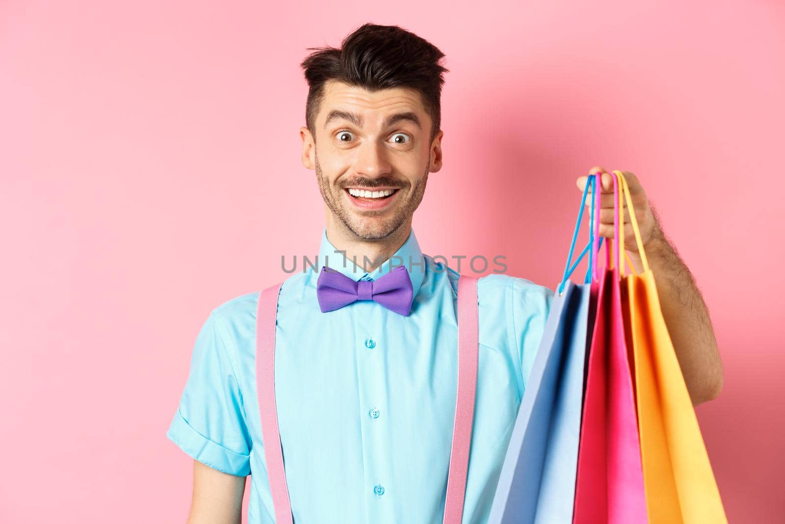 Image of happy guy on shopping, holding paper bags and smiling excited, shopper buying with discounts, standing on pink background.