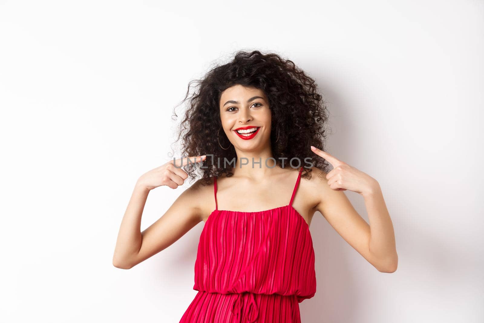 Beautiful woman with curly hairstyle, makeup and red dress, pointing at herself, smiling with white teeth, standing on white background.