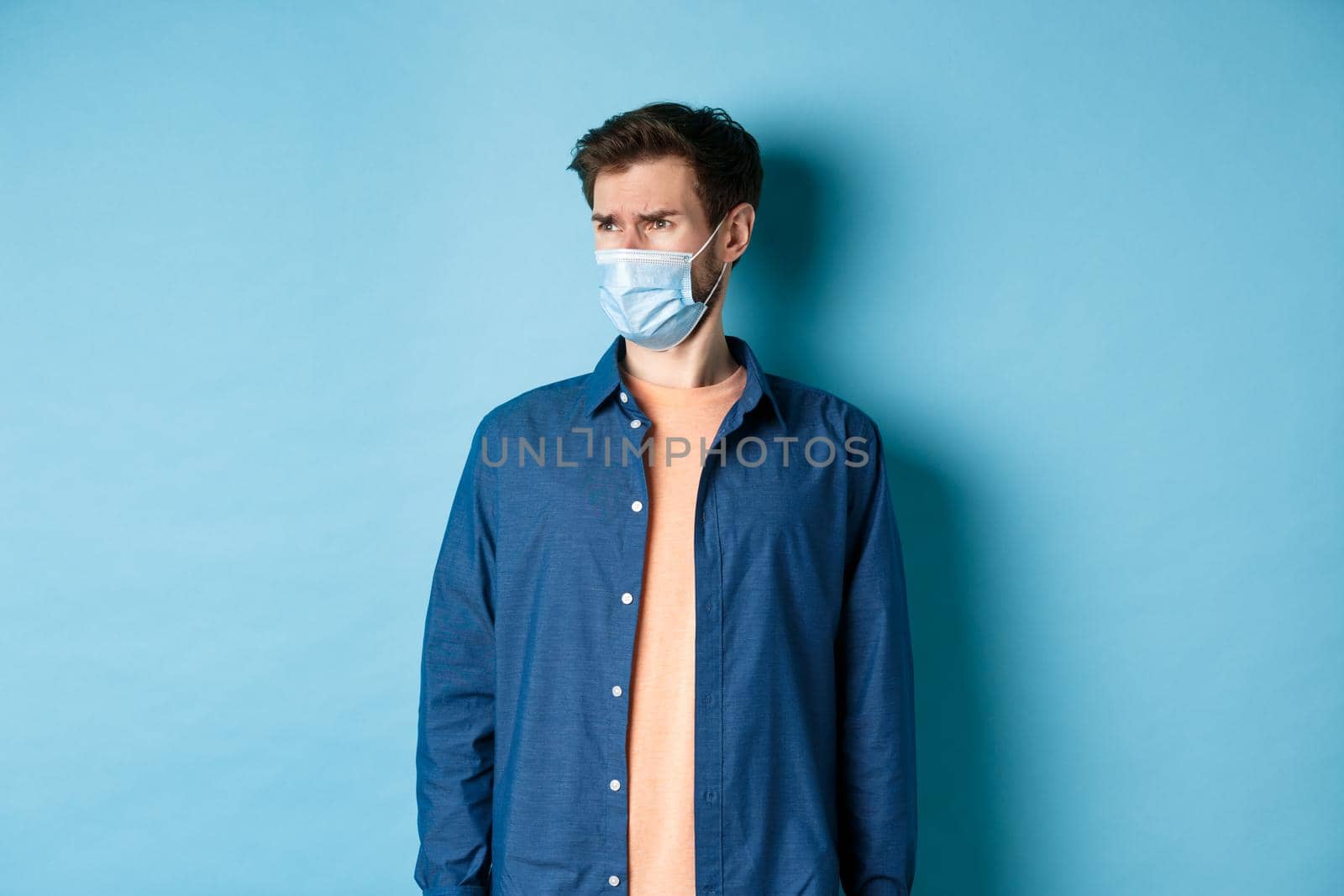 Covid-19 and healthcare concept. Dissatisfied frowning guy in medical mask looking at bad promo, stare aside at empty space with disappointed face, blue background by Benzoix