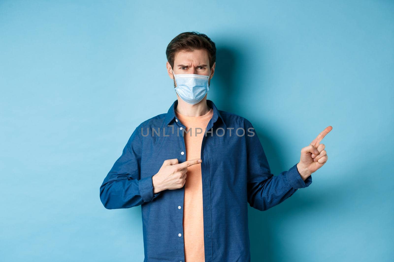 Covid-19 and pandemic concept. Angry man in medical mask frowning, pointig fingers at upper right corner empty space, complaining, standing on blue background by Benzoix