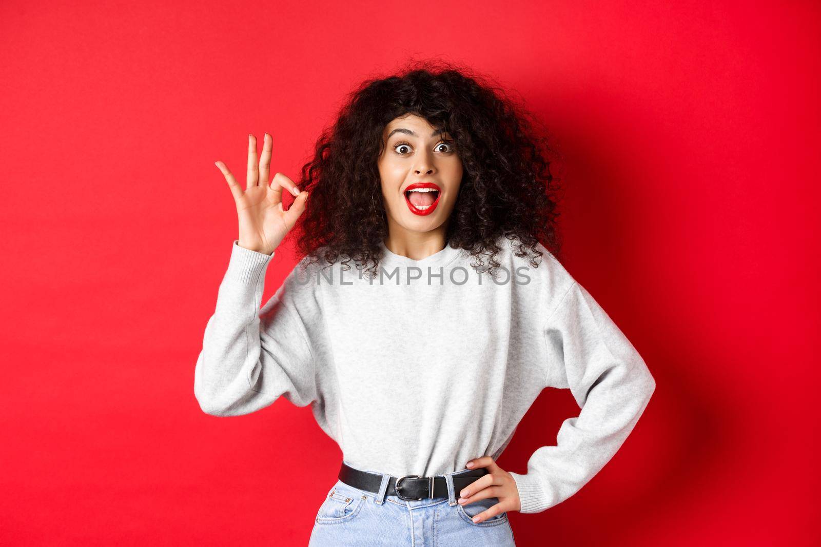 Excited pretty woman say yes, showing okay gesture and looking enthusiastic, agree or like something, making compliment, standing on red background.