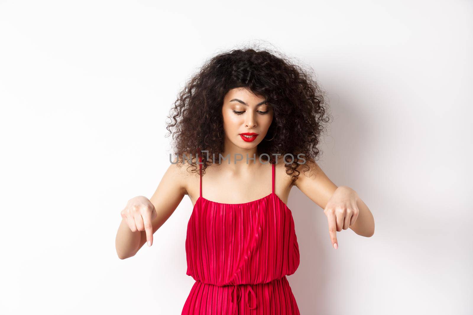 Intrigued beautiful woman with red lips and dress, pointing and looking down at banner, reading logo, standing over white background by Benzoix