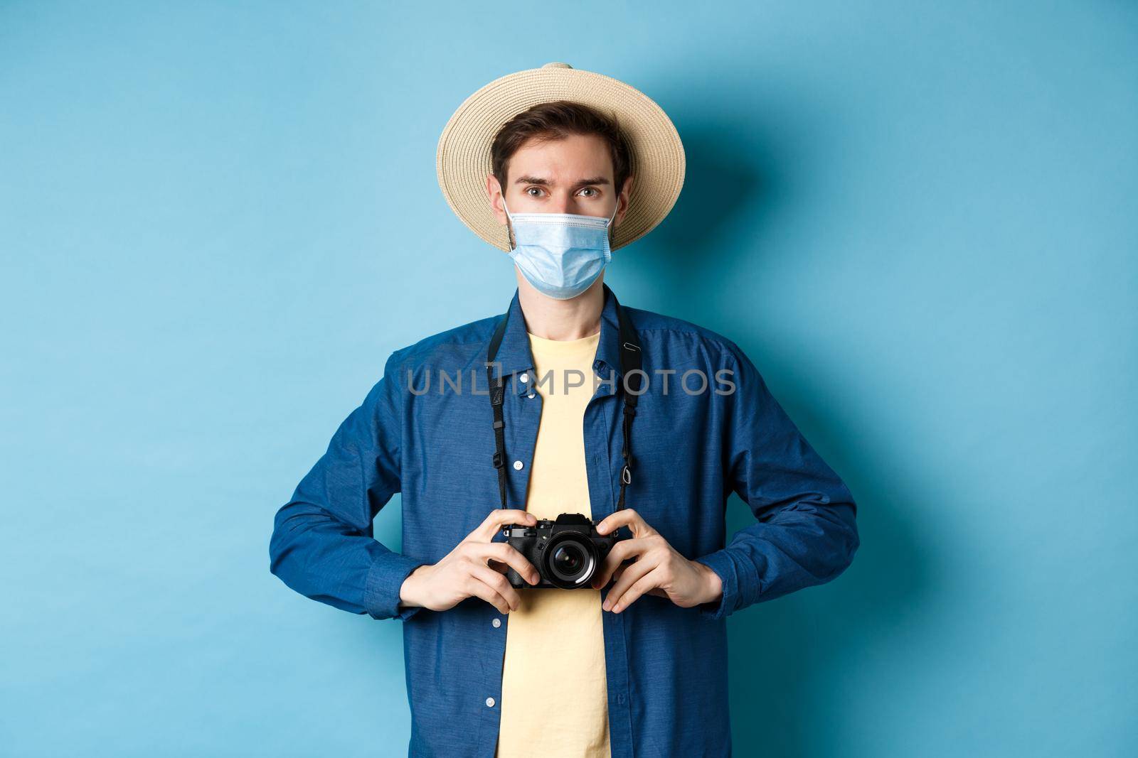 Covid-19, pandemic and travel concept. Young guy travelling abroad with camera, taking pictures on vacation, wearing medical mask from coronavirus, blue background.