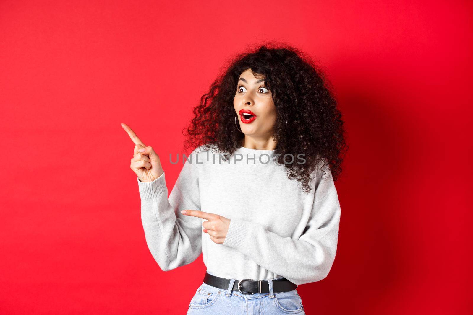 Surprised attractive girl with curly hair, pointing and looking aside at empty space, showing logo or advertisement, standing on red background.