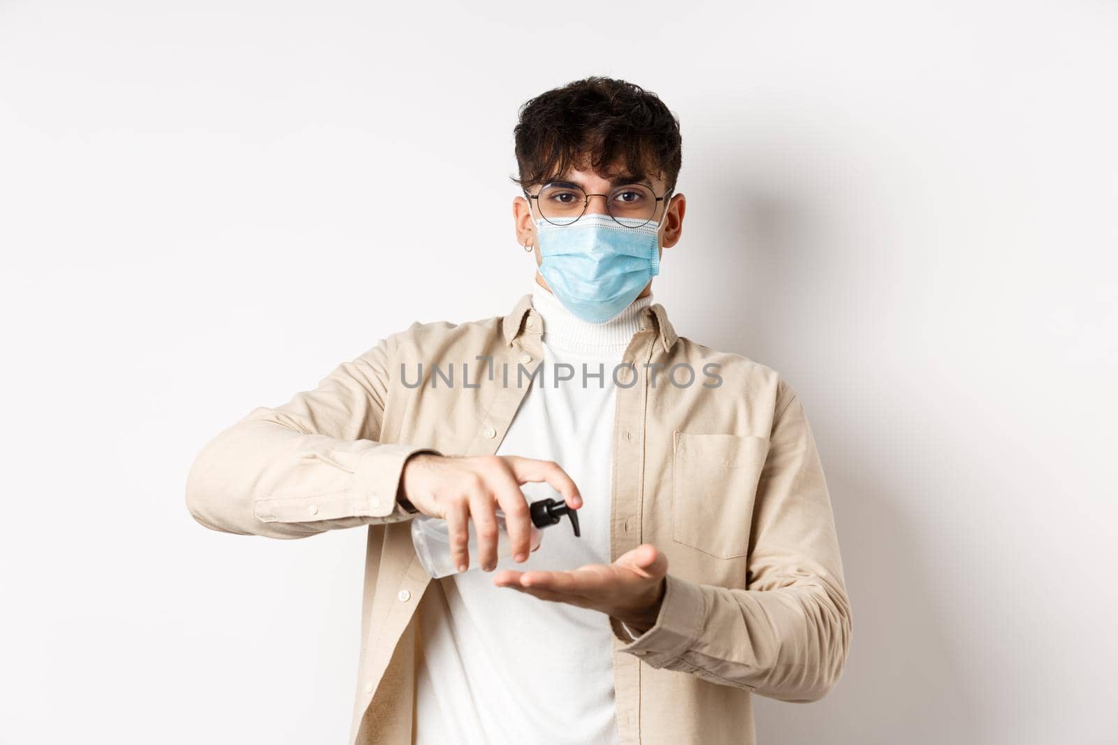 Health, covid and quarantine concept. Young hispanic guy in glasses and face mask using hand sanitizer, apply antiseptic and look at camera, standing on white background.