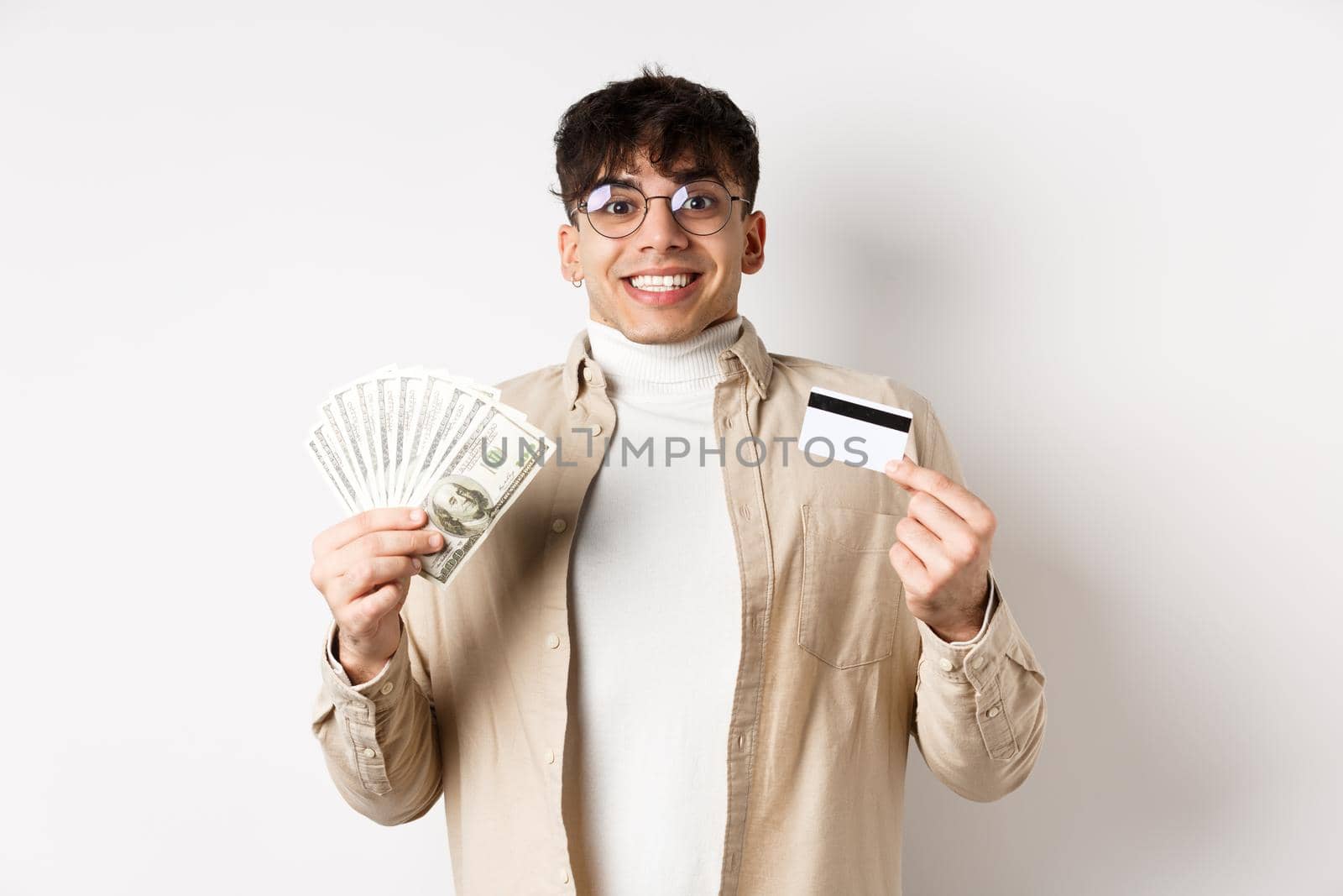 Excited and happy smiling man holding money and plastic credit card, looking amused at camera, standing on white background.