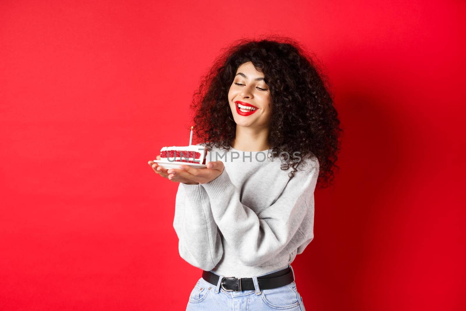 Happy birthday girl celebrating and making wish, holding bday cake and smiling, standing on red background.