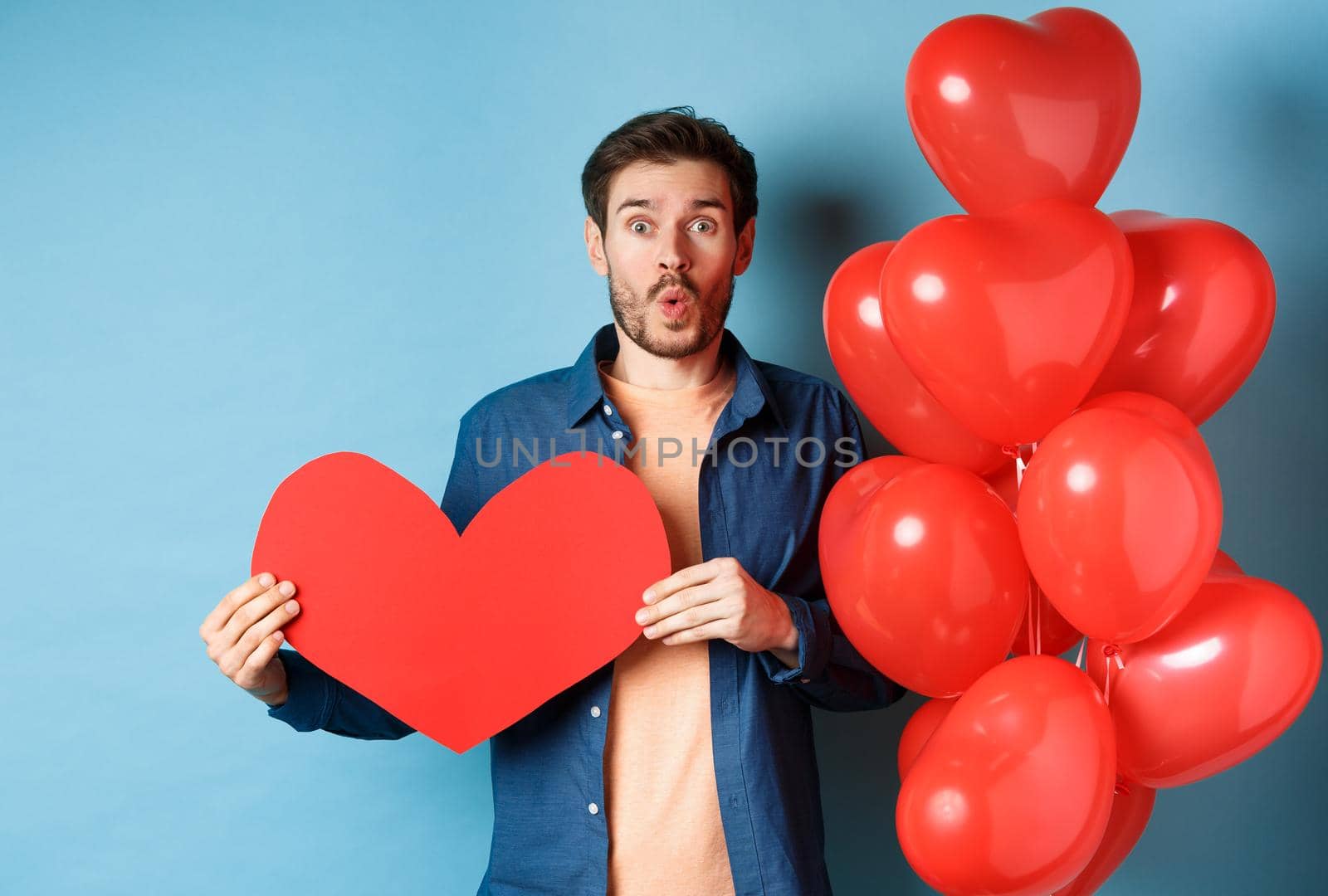 Valentines day concept. Boyfriend in love looking amazed at camera, holding red paper heart and standing with romantic balloon, blue background.