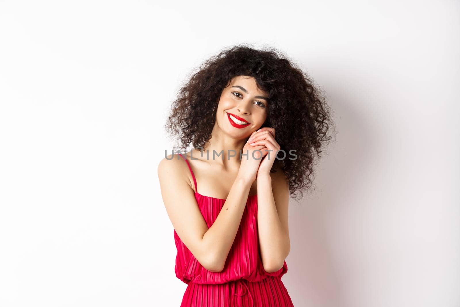 Lovely young woman with curly hairstyle, wearing red dress, looking at silly and tender thing, admire something, standing over white background by Benzoix