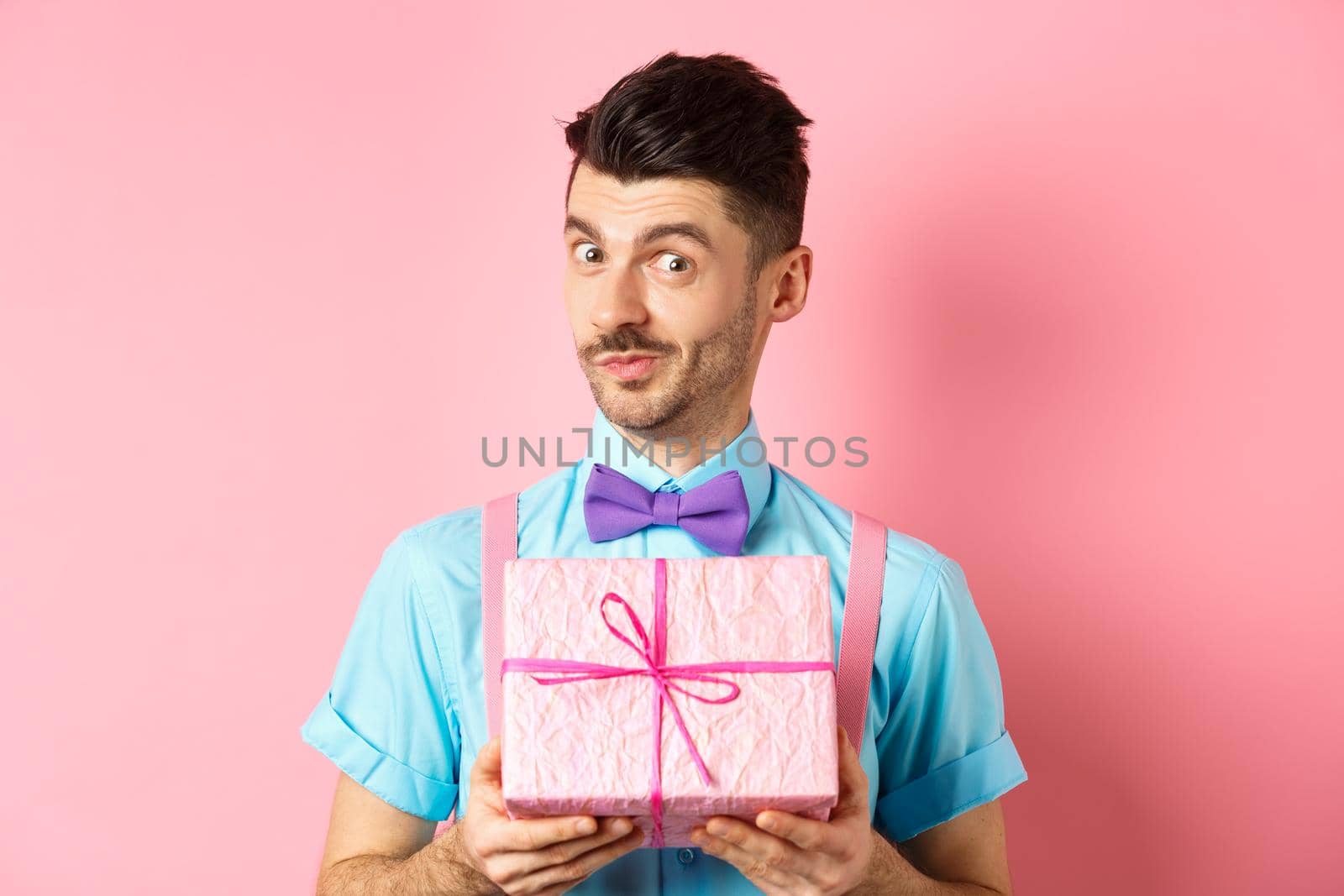 Holidays and celebration concept. Funny guy with moustache showing cute gift box wrapped in festive paper, looking at camera, presenting you, standing on pink background.