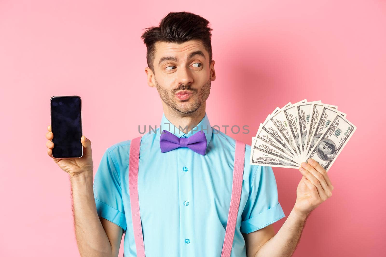 E-commerce and shopping concept. Thoughtful young man in bow-tie showing empty smartphone screen, looking pensive at money, standing on pink background.