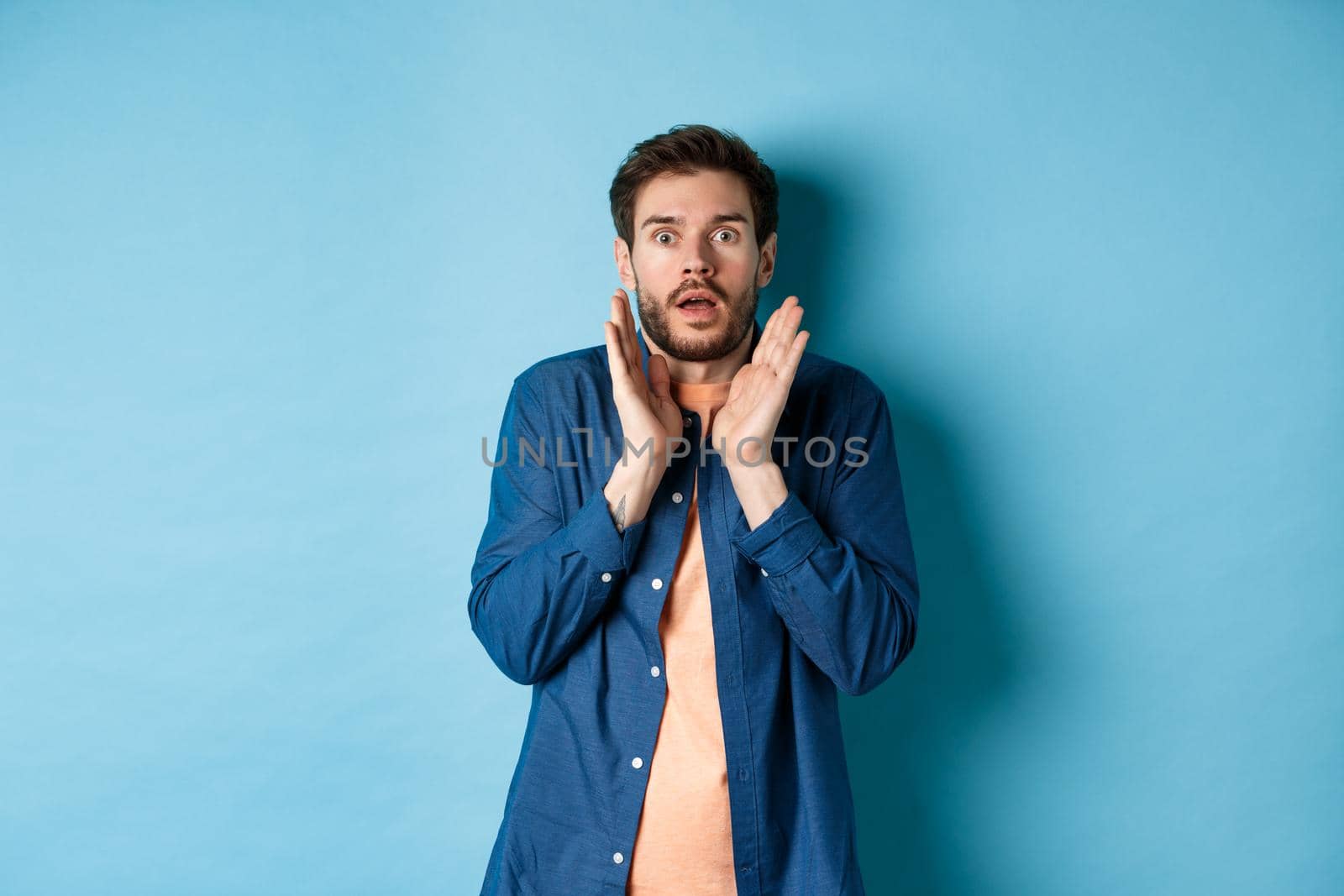 Shocked and scared young man standing in stupor, raising hands up and gasping speechless, standing on blue background.