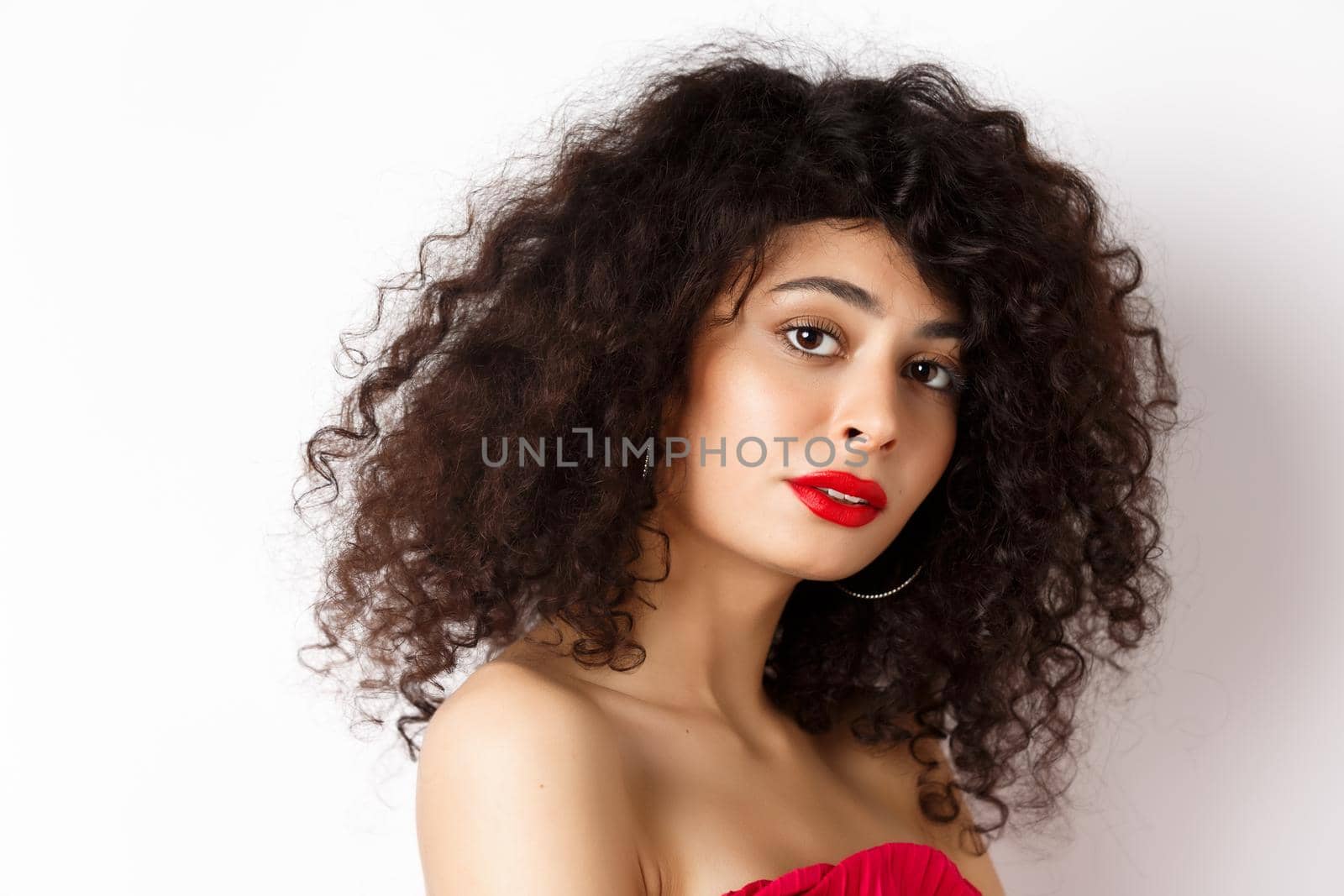 Close-up of beautiful caucasian woman with curly haircut and red lipstick, looking tender at camera, standing over white background.