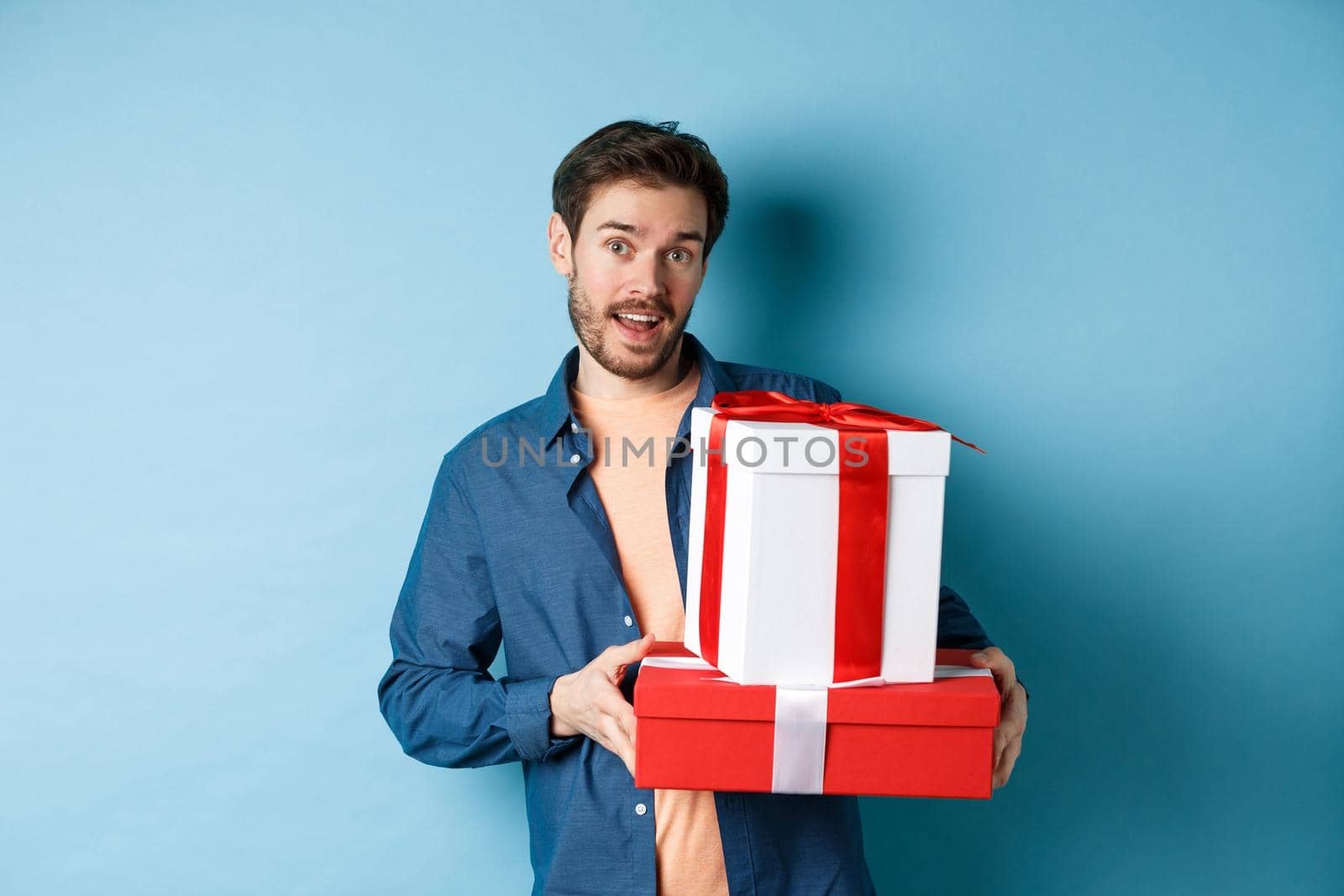 Handsome bearded guy holding Valentines day gifts for lover, standing with presents in boxes and looking at camera, blue background.