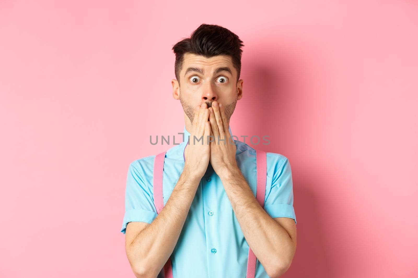 Shocked man looking in startled, covering mouth with hands and staring at camera, standing on pink background.