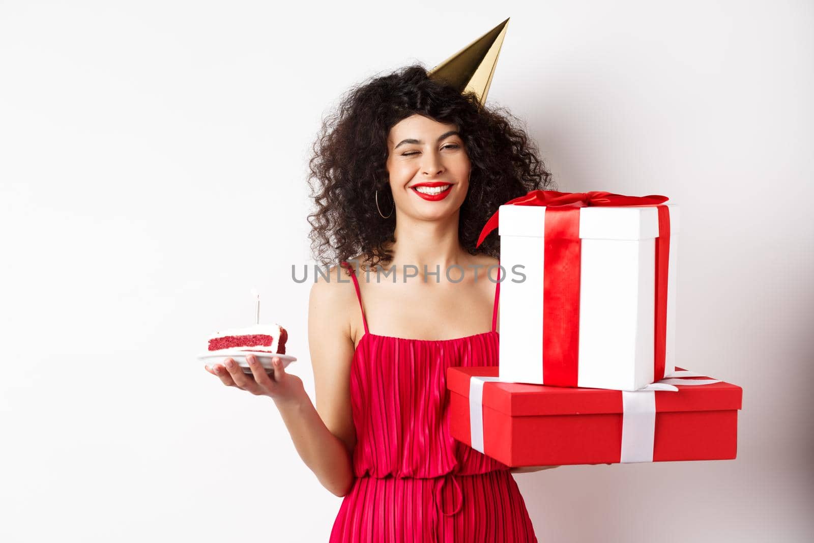 Happy birthday girl in red dress, celebrating and holding gifts with bday cake, standing on white background.