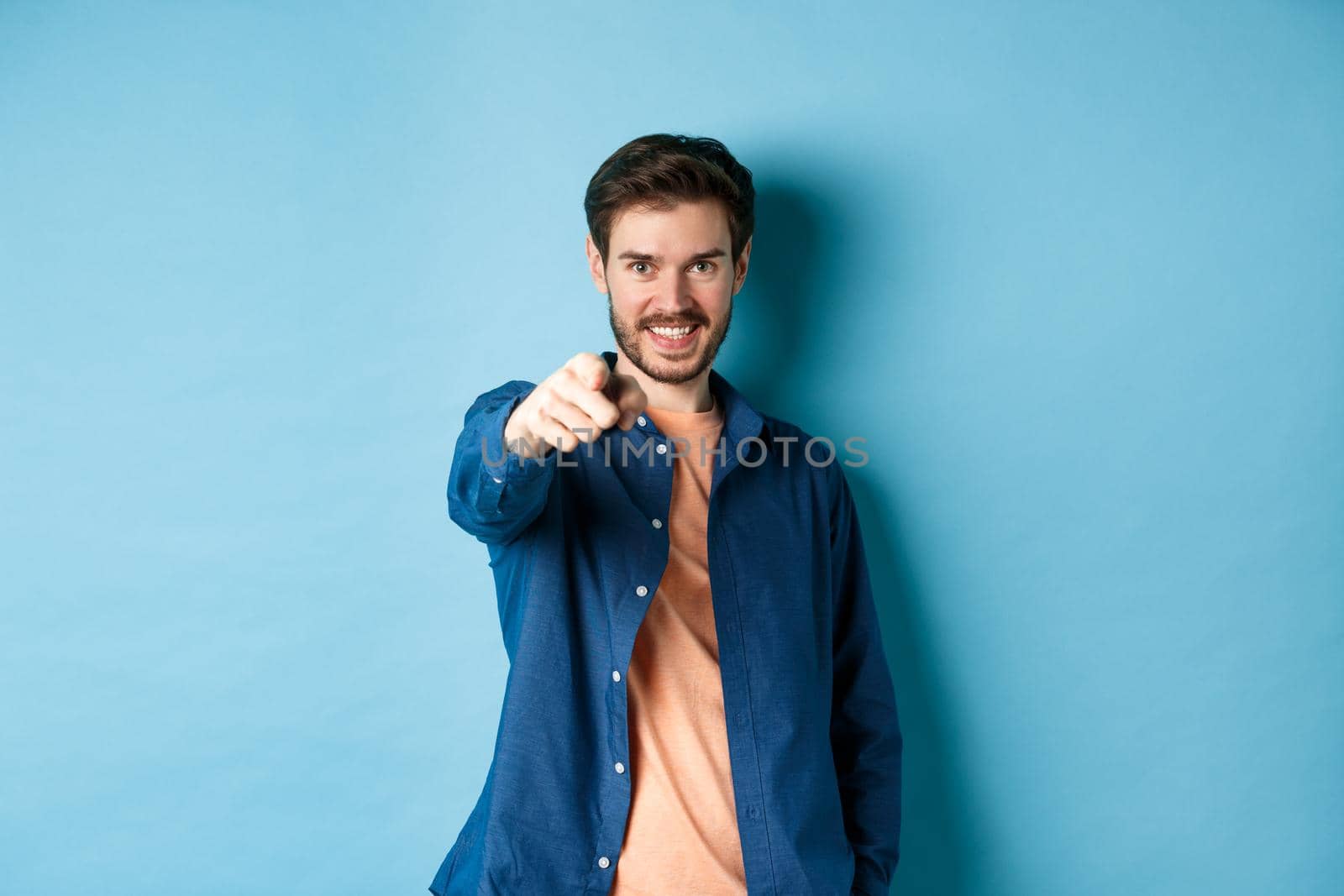 Confident smiling guy choosing or inviting you, pointing finger at camera decisive, standing on blue background by Benzoix