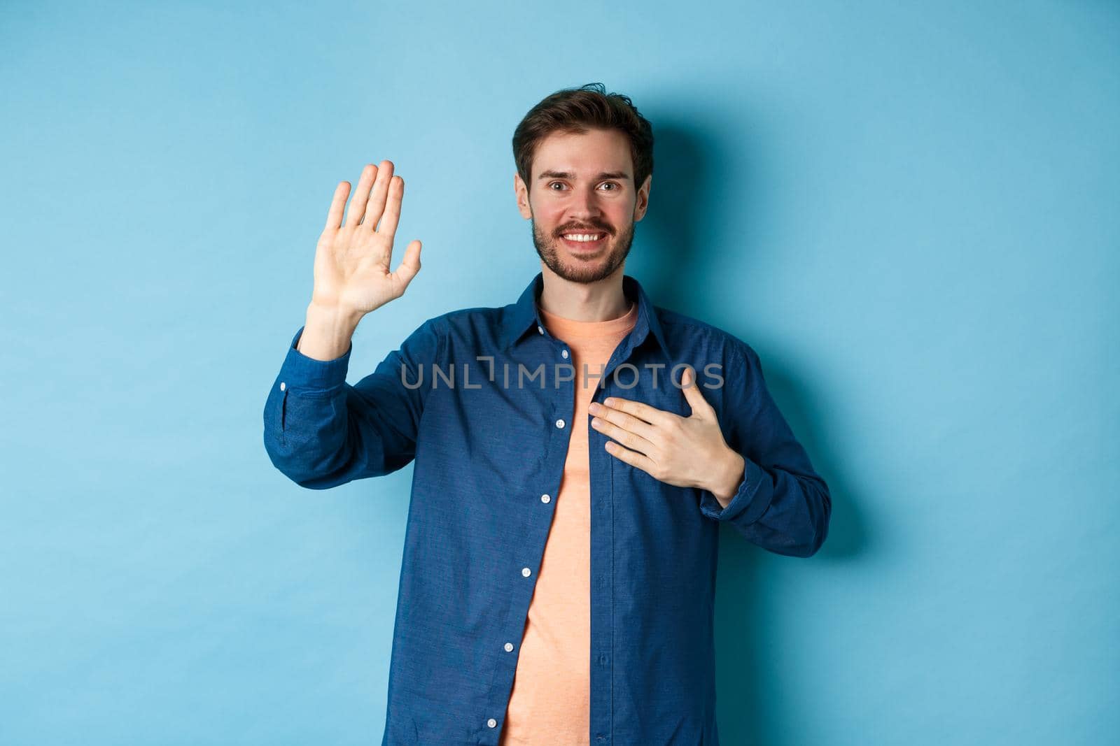 Happy young man introduce himself, telling truth with hand on heart and friendly smile, standing on blue background.
