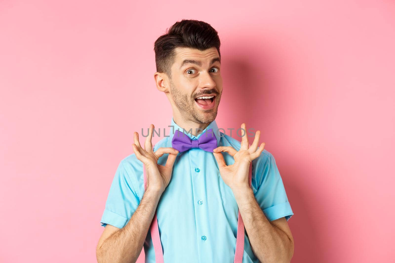 Classy handsome man fixing bow-tie on neck and smiling, getting ready for date or party, standing on pink background.