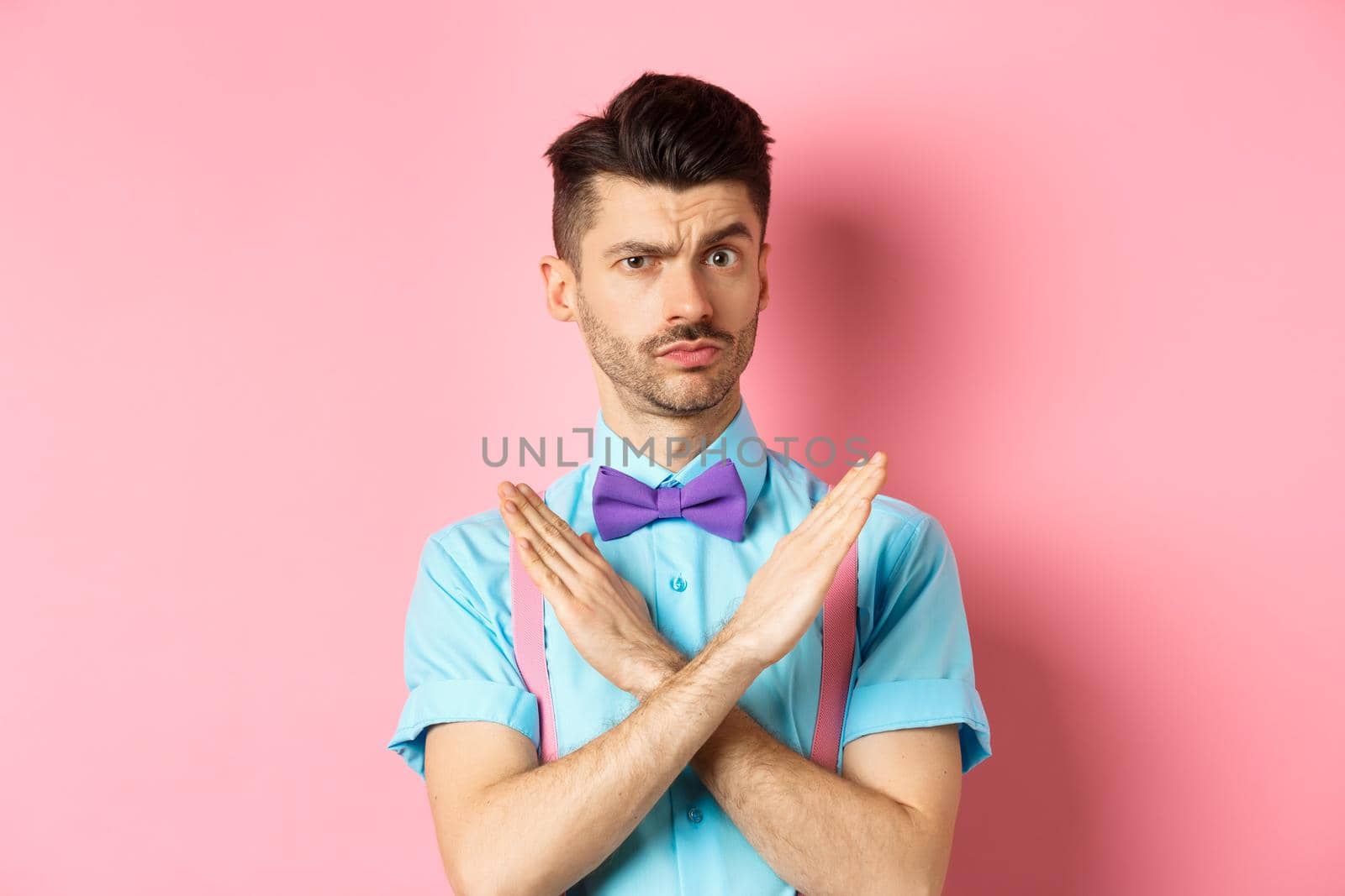 Serious-looking skeptical guy showing cross sign to stop, prohibit something bad, forbid action, standing on pink background by Benzoix