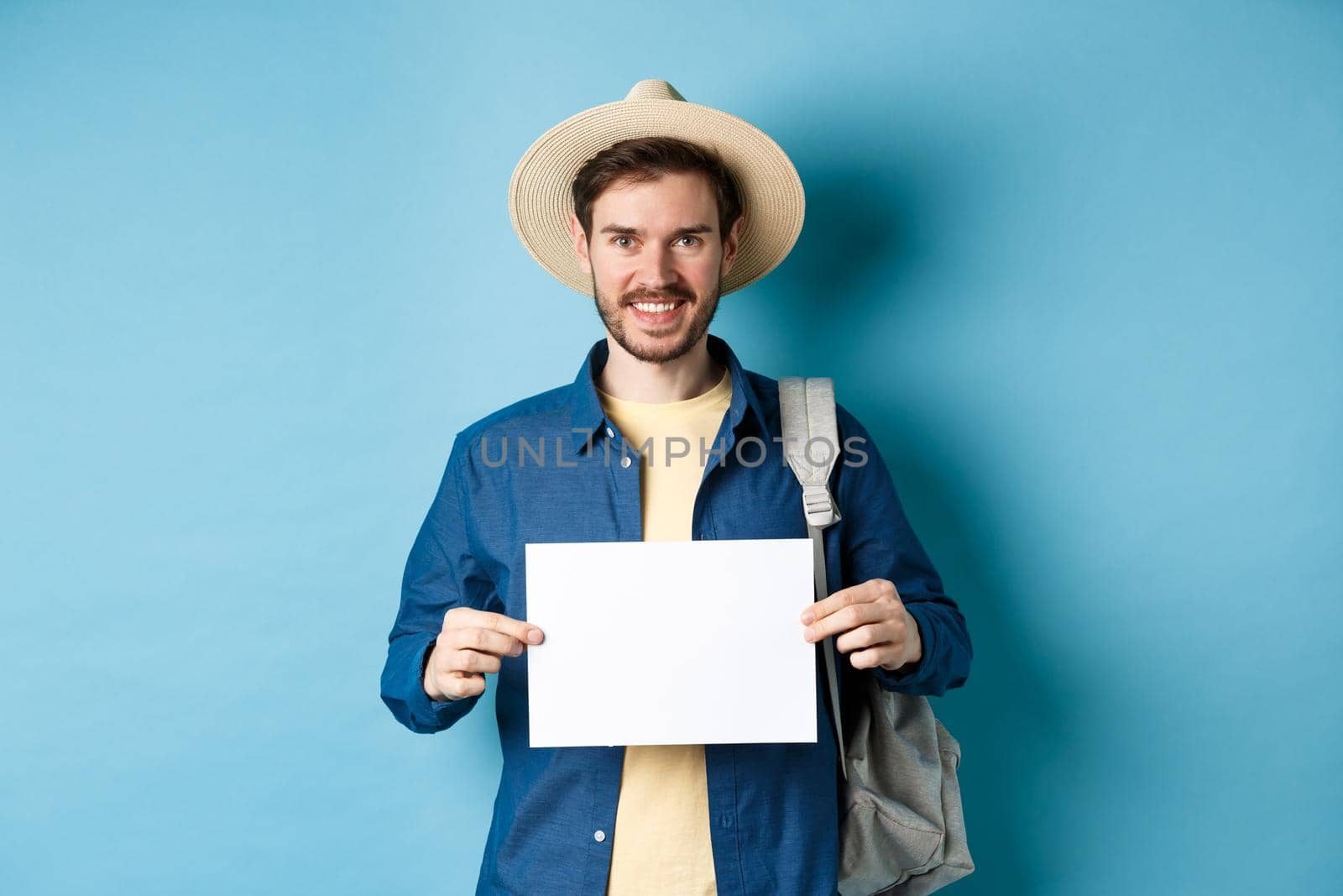 Happy tourist with backpack showing empty piece of paper, smiling at camera, standing on blue background. Concept of summer holiday and vacation by Benzoix
