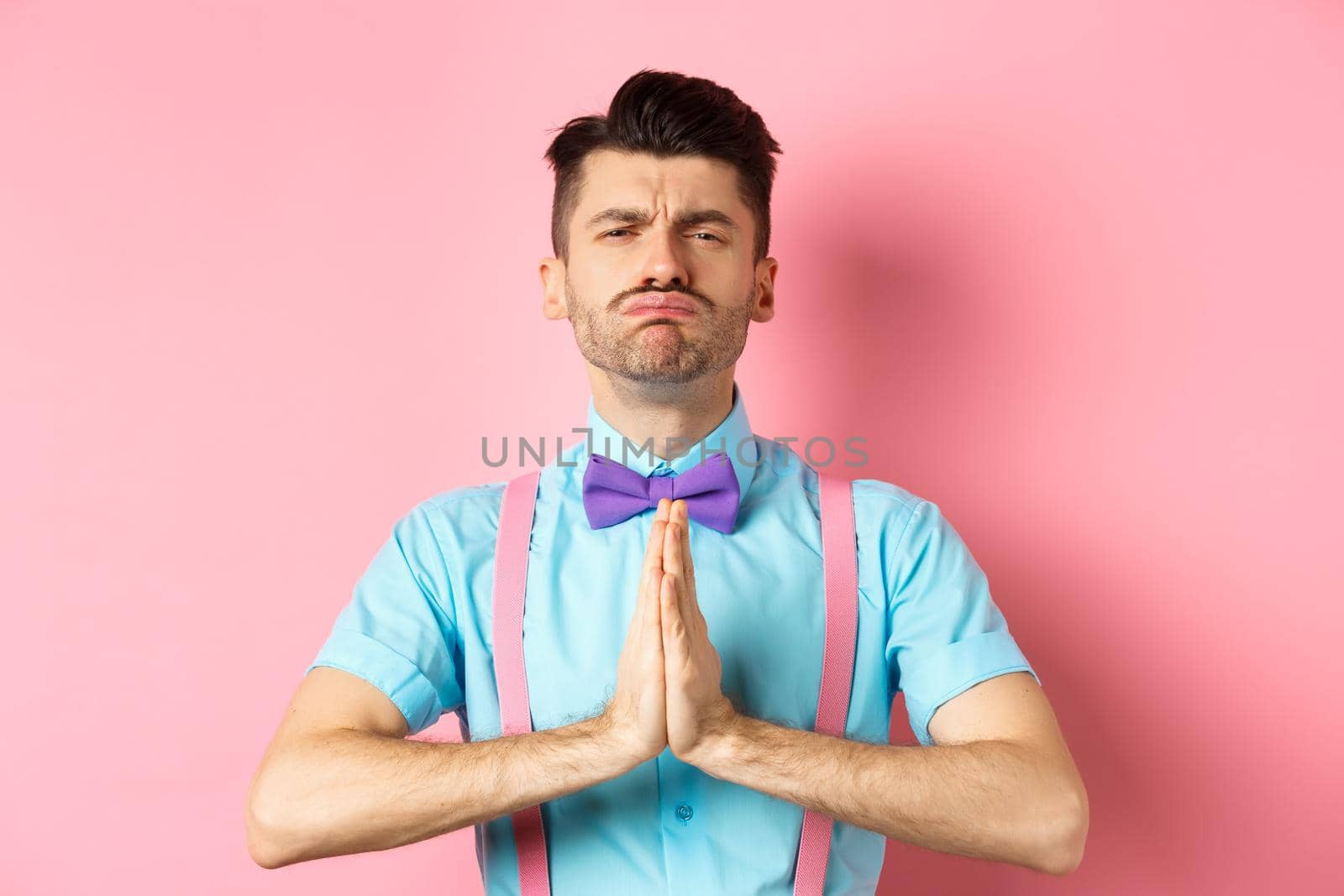 Silly guy making begging face, pleading and asking for help, standing with hands pressed together on pink background. Copy space
