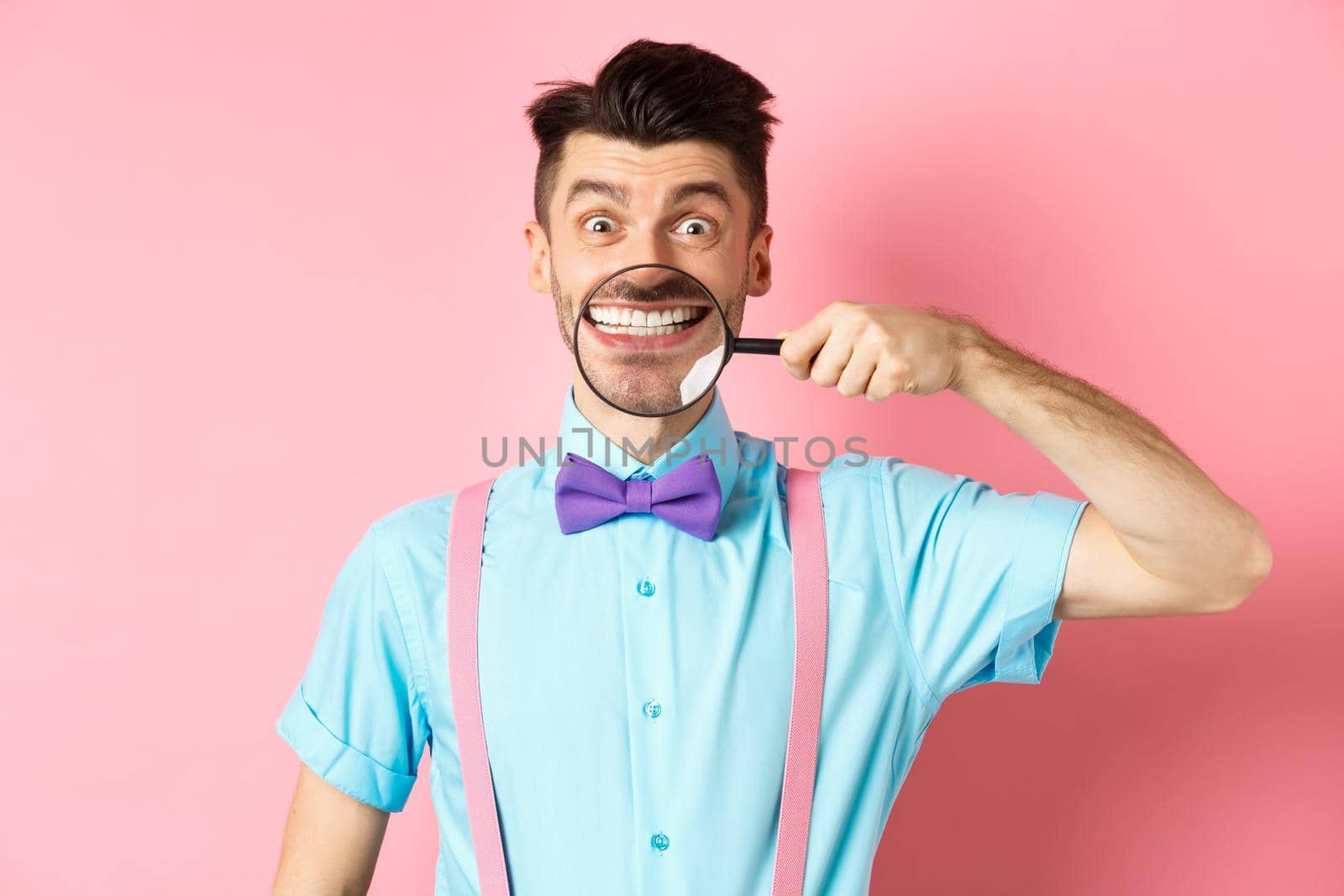 Funny caucasian guy in bow-tie showing his white smile teeth with magnifying glass, looking cheerful at camera, standing on pink background.