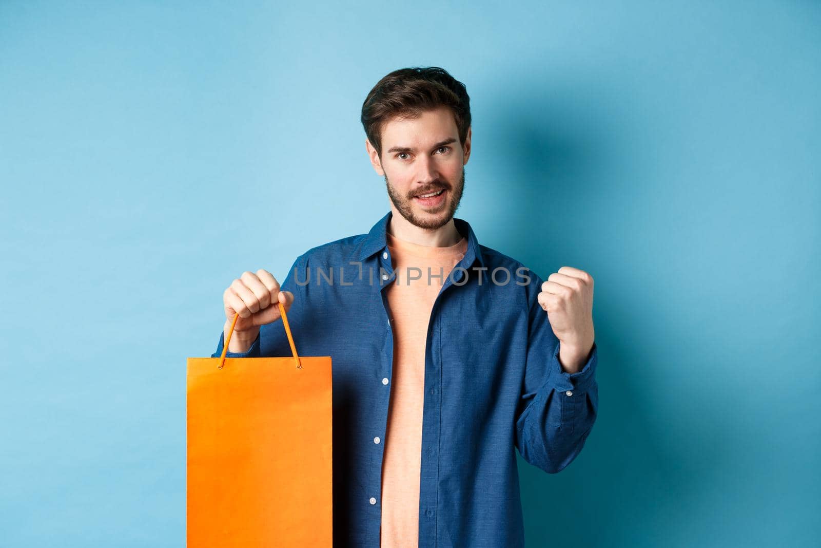 Handsome smiling man saying yes, triumphing and showing shopping bag, celebrate good promo offer, standing on blue background by Benzoix