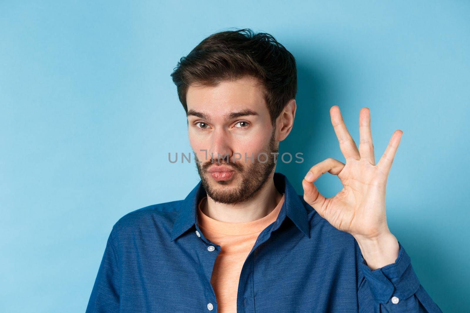 Image of silly young man pucker lips and show okay gesture, praise good thing, compliment excellent choice, standing on blue background by Benzoix