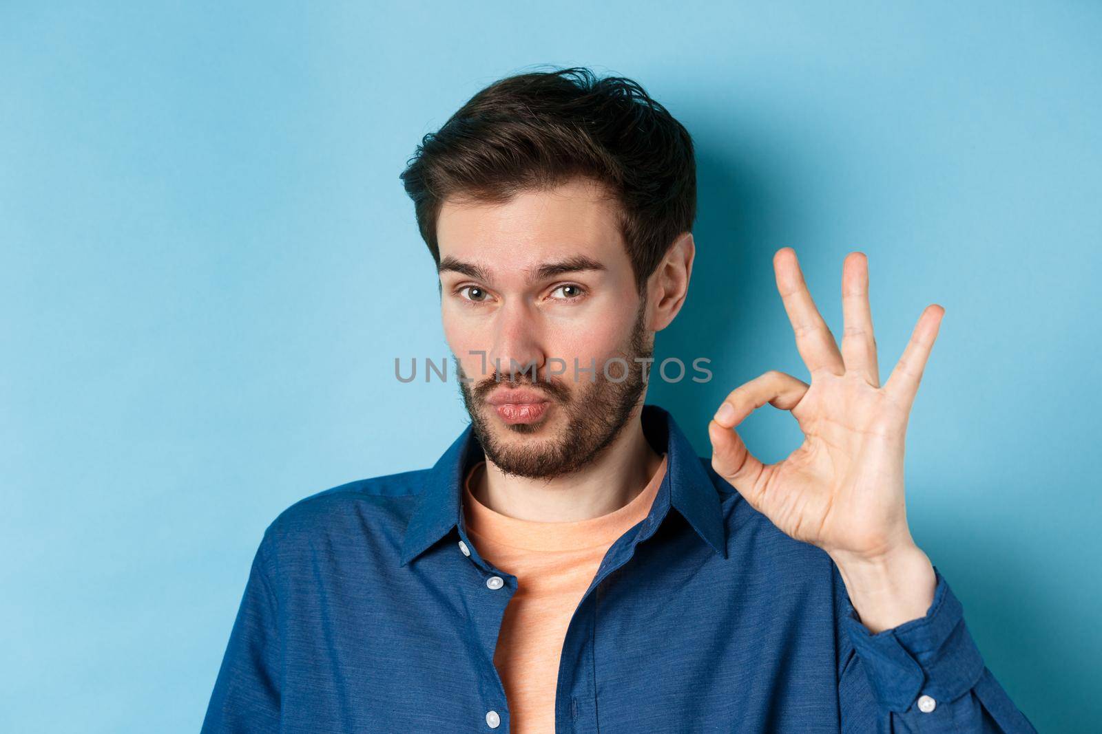 Image of silly young man pucker lips and show okay gesture, praise good thing, compliment excellent choice, standing on blue background.