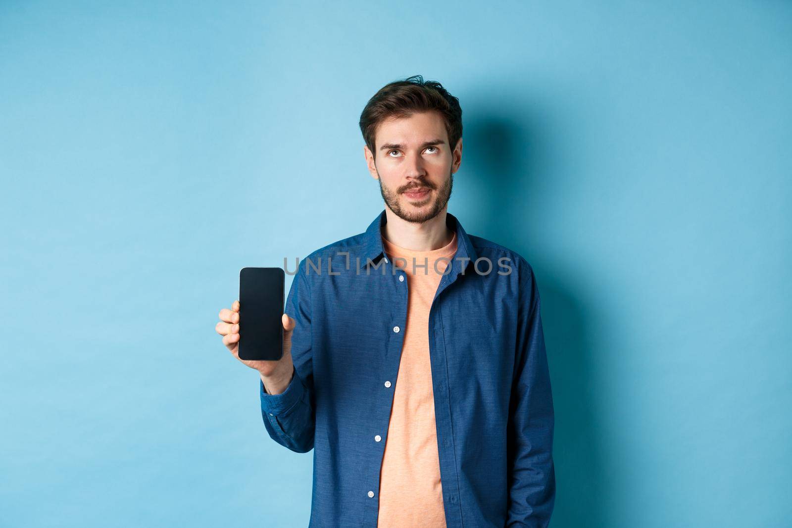 Annoyed young guy showing empty smartphone screen and roll eyes up bothered, standing on blue background by Benzoix