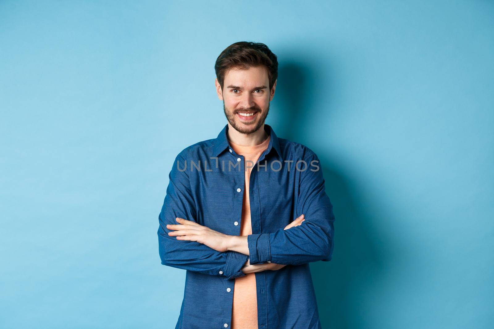 Confident young man feeling like professional, cross arms on chest and smiling sassy, standing on blue background by Benzoix
