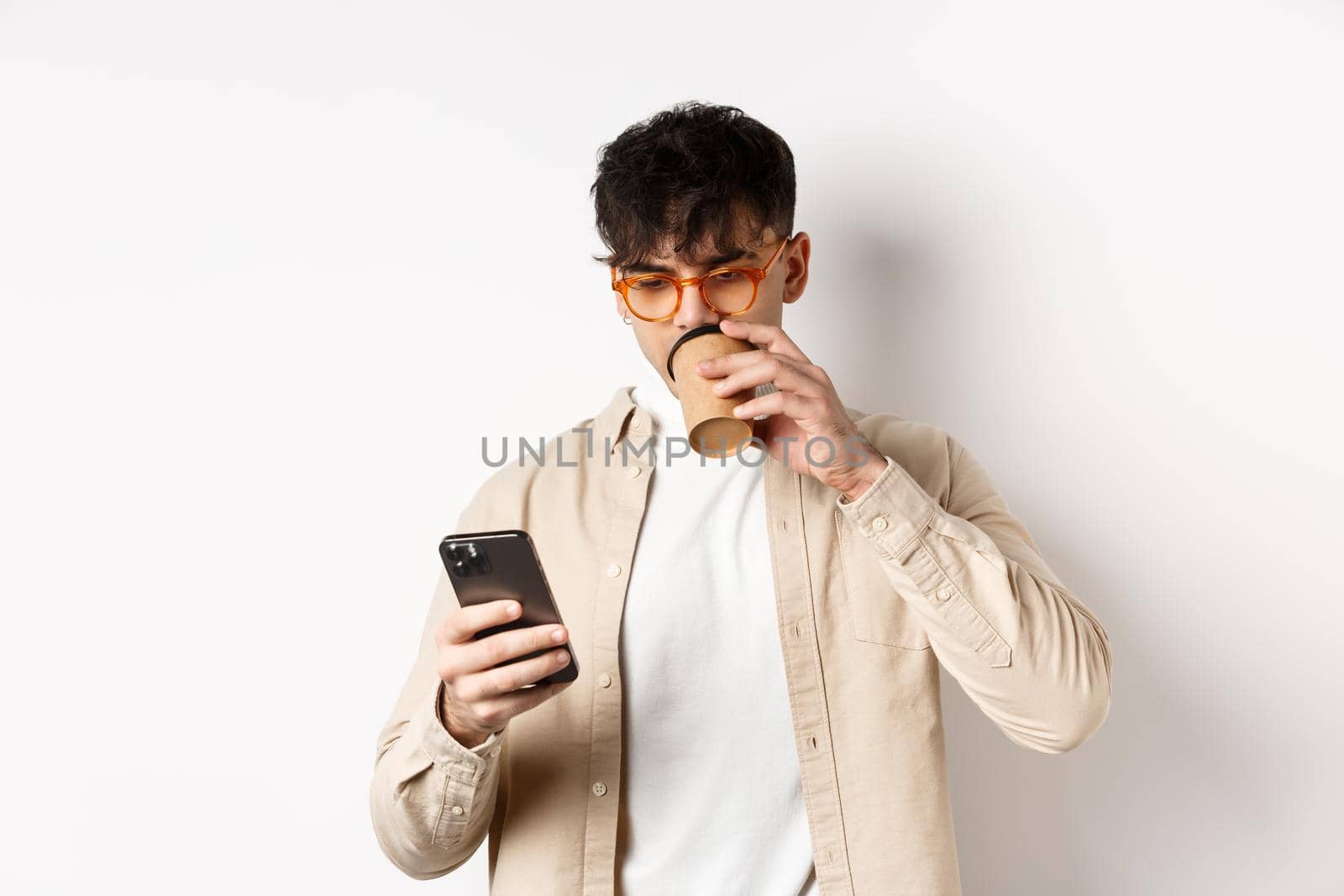 Real people. Young man reading smartphone screen and drinking coffee, looking at phone, standing on white background.