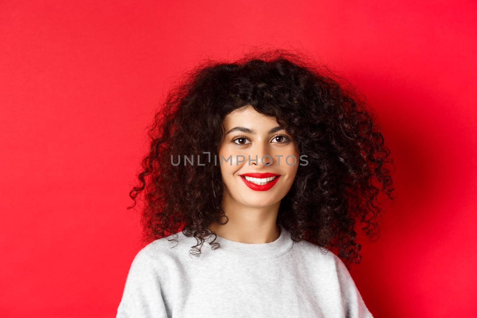 Close-up of beautiful lady with red lips and curly hair, smiling and looking happy at camera, studio background.